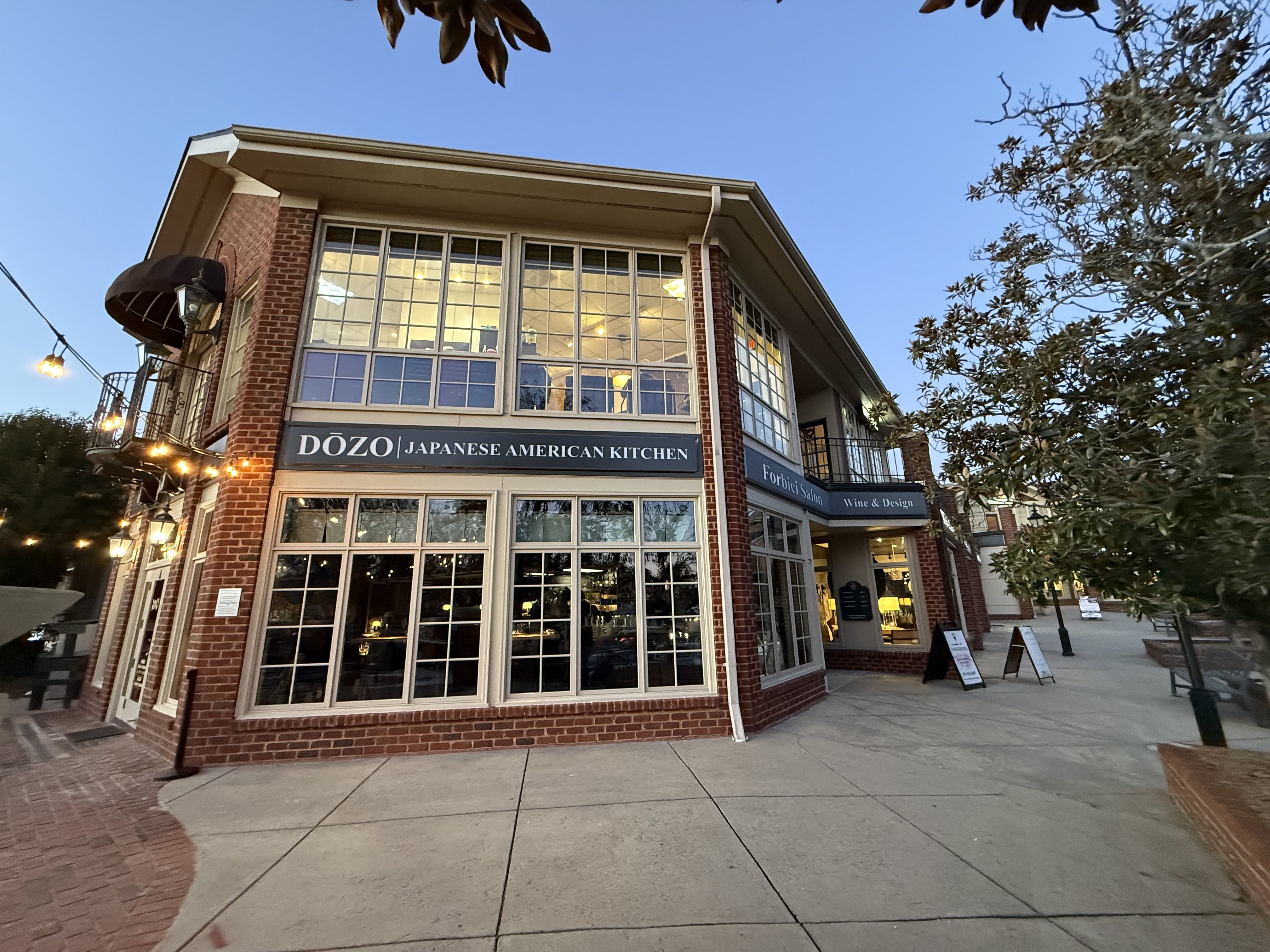 Brick building with large windows under a clear sky, featuring "Dōzo Japanese American Kitchen" and "Forbici Salon Wine & Design" signs, with lit outdoor string lights and nearby trees.