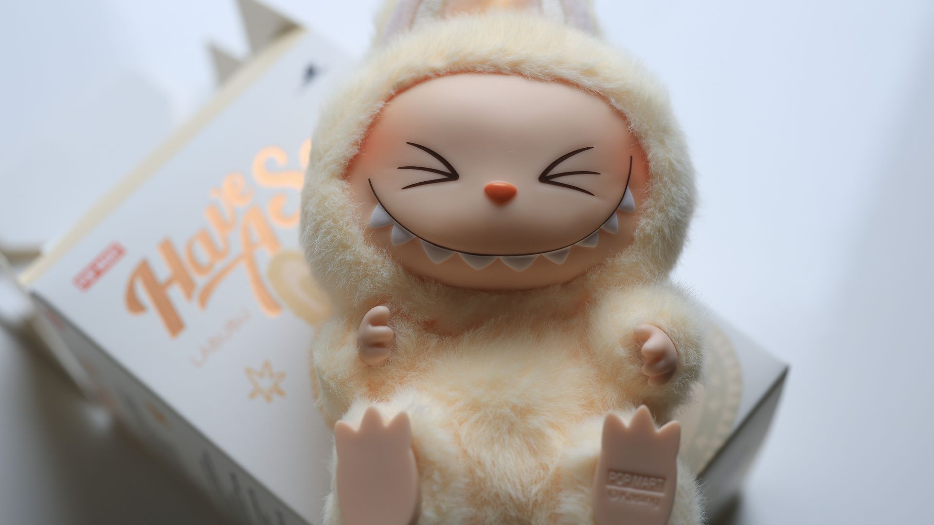 Soft, beige plush toy wearing a fuzzy costume with ears; it has a cute, closed-eye, toothy smile and an orange nose, sitting inside a pale box.