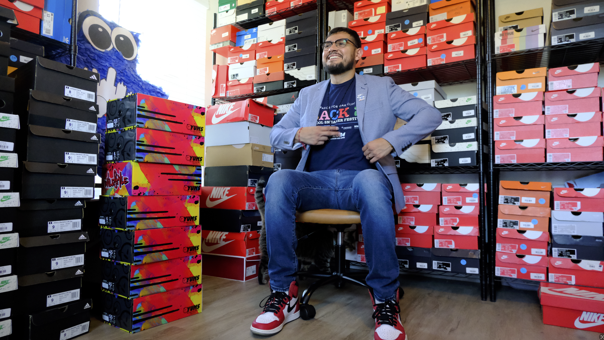 Man in glasses, blue blazer, and red-white-black Nike sneakers sits on a chair surrounded by stacks of colorful shoe boxes in a room filled with sneakers.