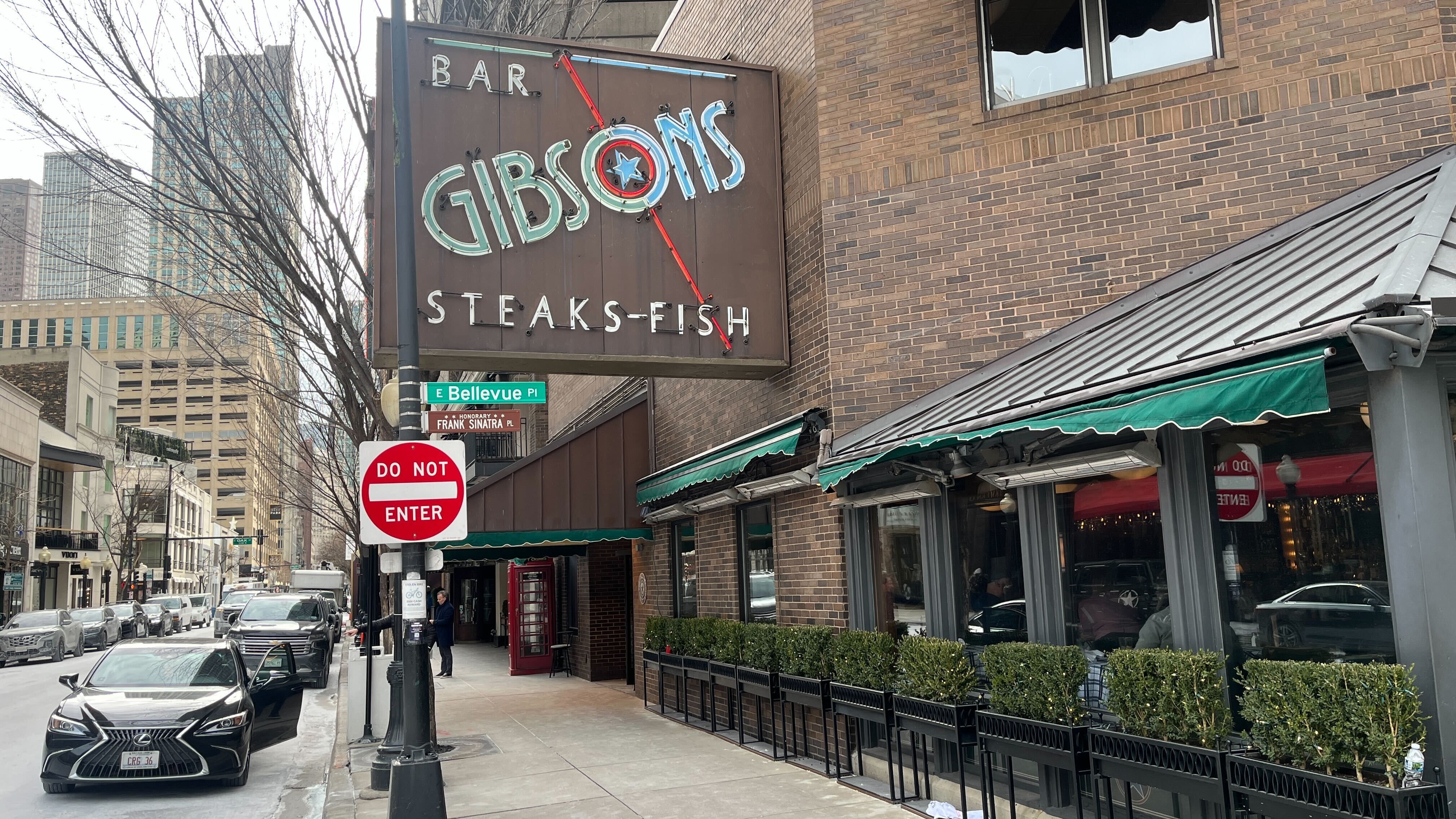 Street view of a brick building with a large sign reading "Bar Gibsons Steaks-Fish," green awnings, a row of planter boxes, a parked Lexus car, and a red "Do Not Enter" sign.