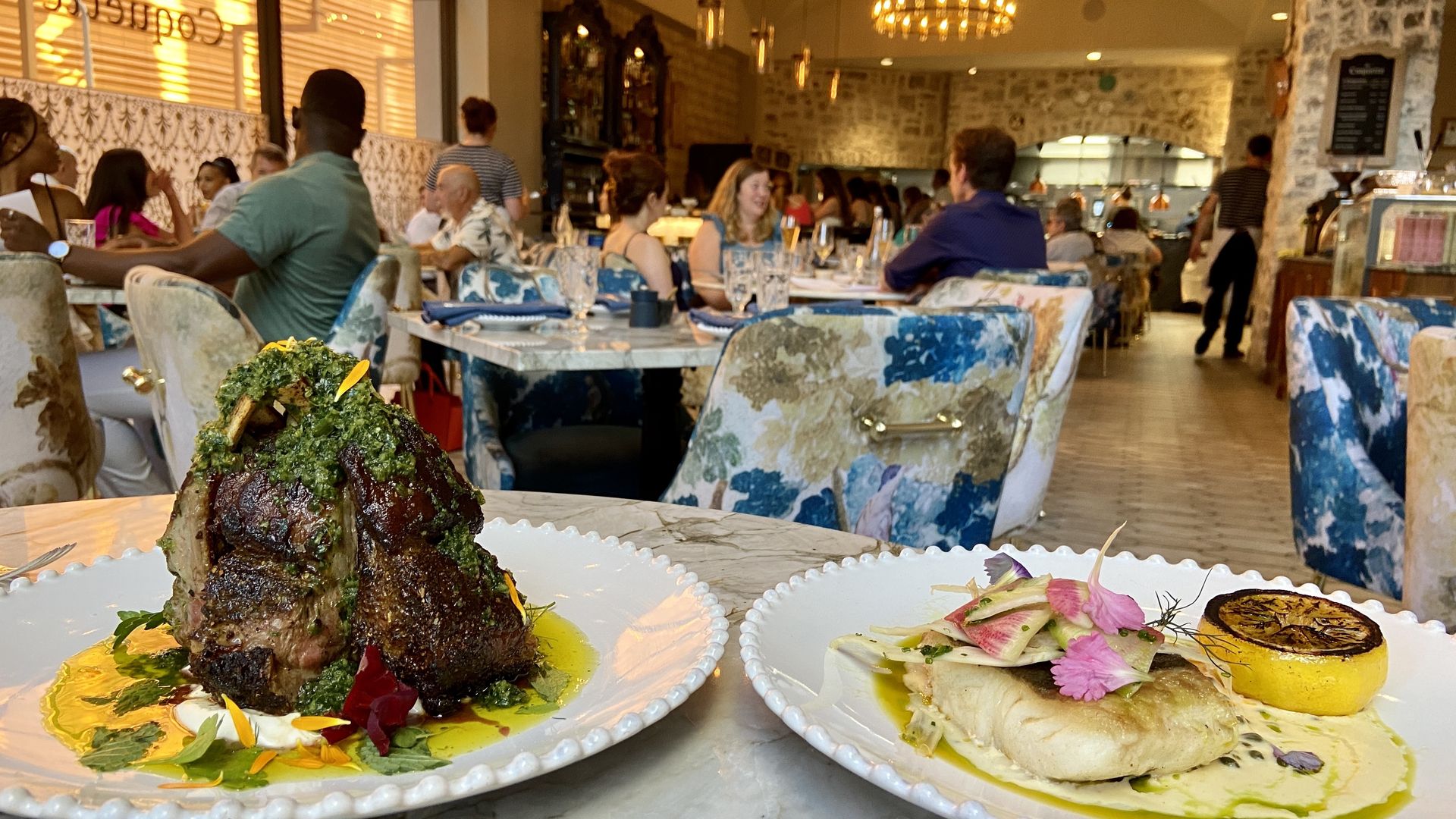 Two gourmet dishes on white plates on a marble table in a lively restaurant; one has lamb chops with green herbs, the other fish with grilled lemon and flower garnish. Patrons chat in background.