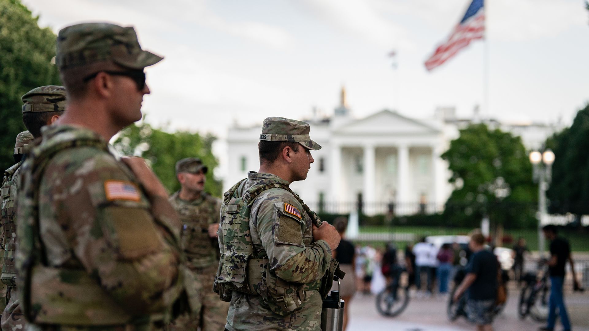 U.S. soldiers in camouflage uniforms stand guard near the White House with an American flag flying above, as people walk and gather nearby on a paved area.