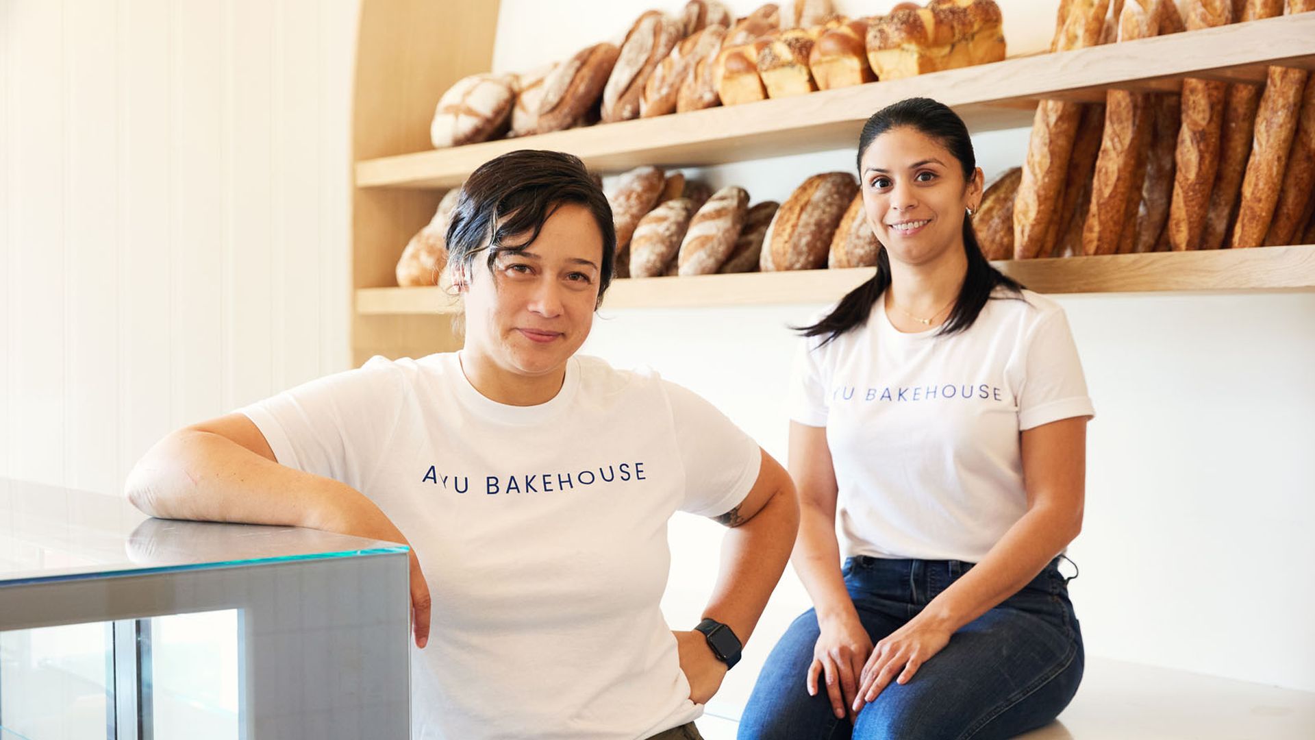 Samantha Weiss and Kelly Jacques pose for a photo at Ayu Bakehouse, each wearing a business T-shirt and with bread stacked in the background.