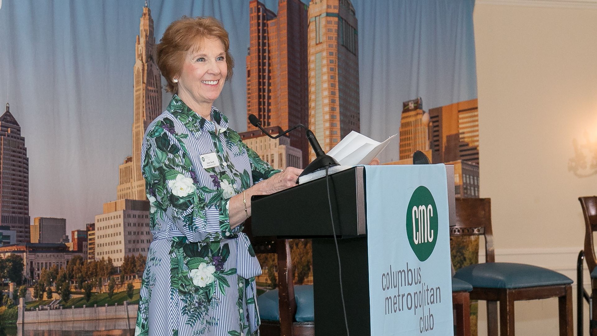 Jane Scott speaks at a podium that reads "Columbus Metropolitan Club" in front of a Columbus skyline backdrop