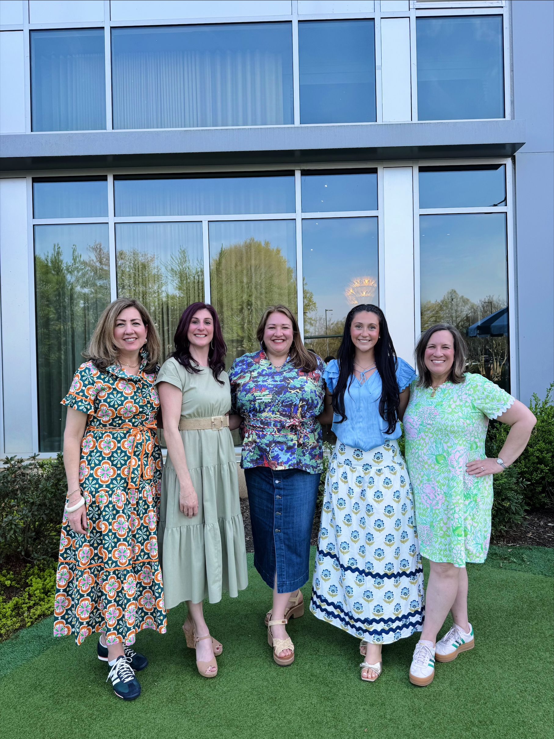 Five women stand on a grassy area in front of a modern glass building, smiling. They wear bright, patterned dresses in orange, sage green, blue, and lime tones.