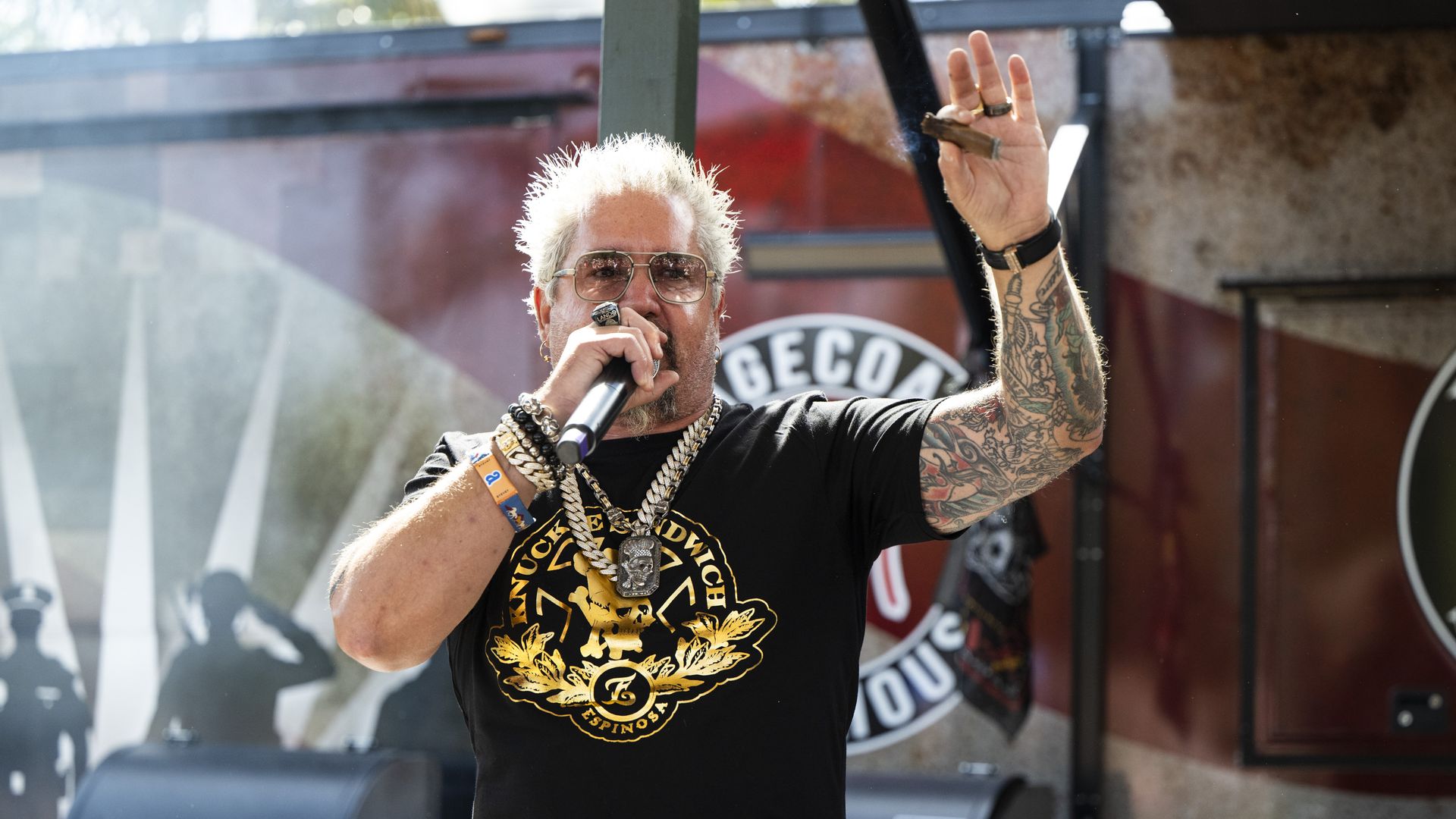 Tattooed man with bleached hair and glasses performs on a stage, wearing a black T-shirt with a gold emblem and heavy chains, holding a microphone and raising a smoking cigar-like item.