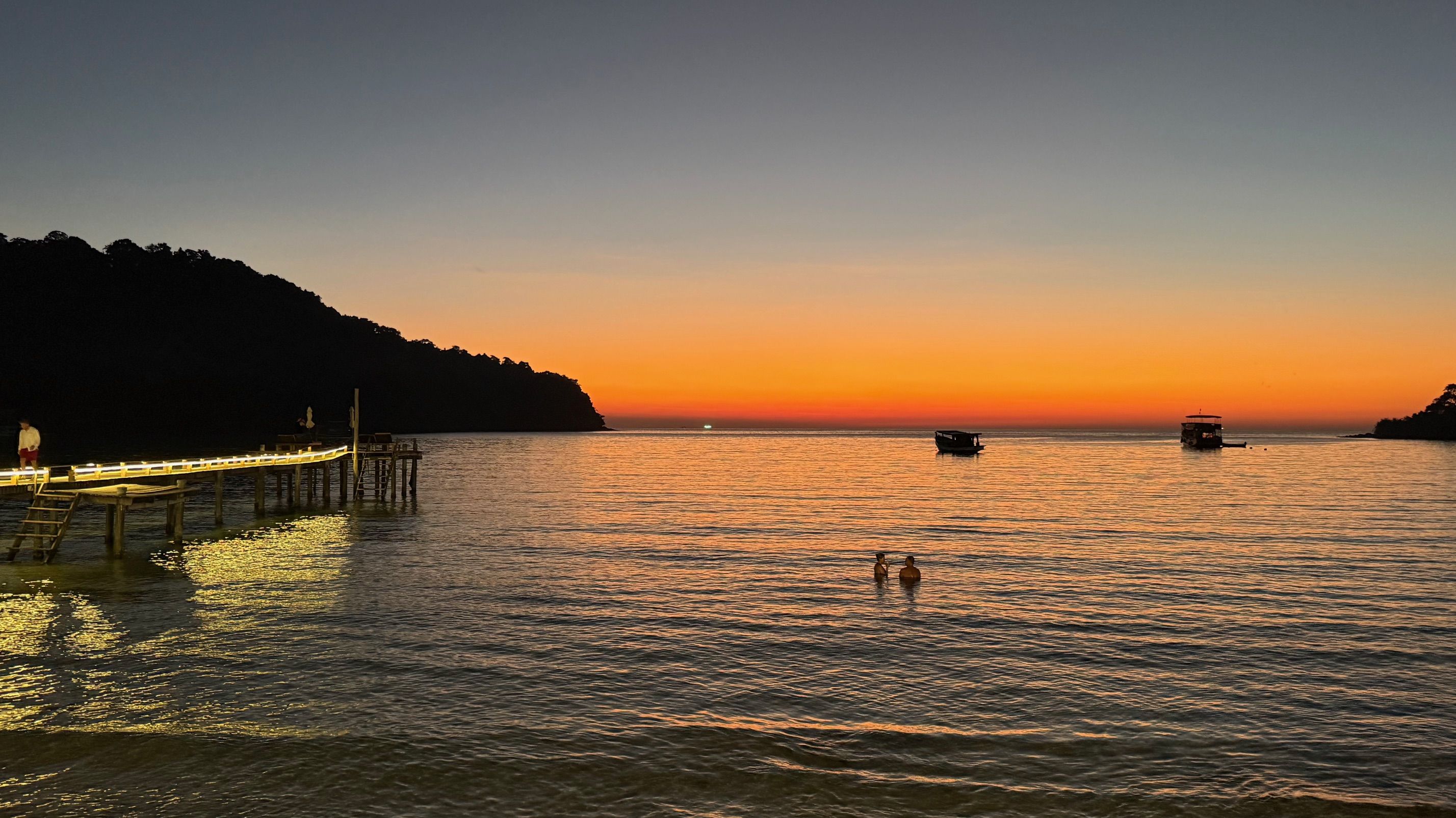 Orange and blue sunset over calm sea with a lit wooden pier, two boats floating, and two people wading in water near the shore facing the horizon.