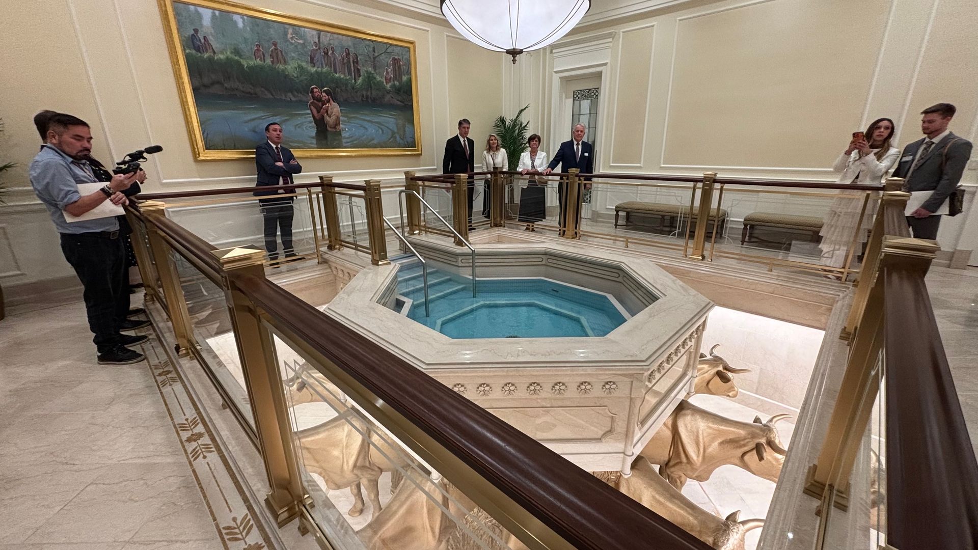 Opulent atrium with a central octagonal marble pool, gold railing, and a large framed painting of a baptism. Several formally dressed people and a camera crew stand around, some taking photos.