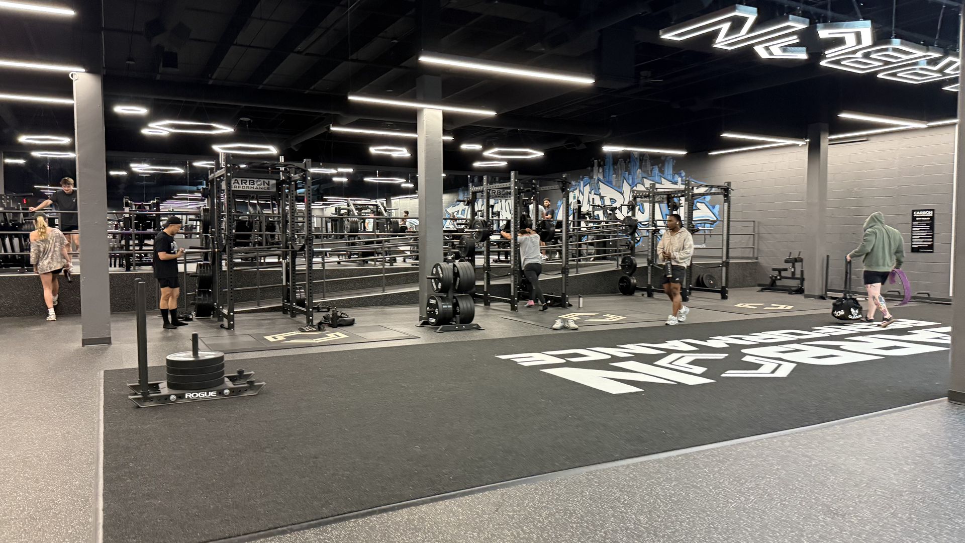 Indoor gym with squat racks and free weights; several people lifting or walking around. Gray walls, black ceiling, bright geometric neon lights overhead, and large white floor graphics.