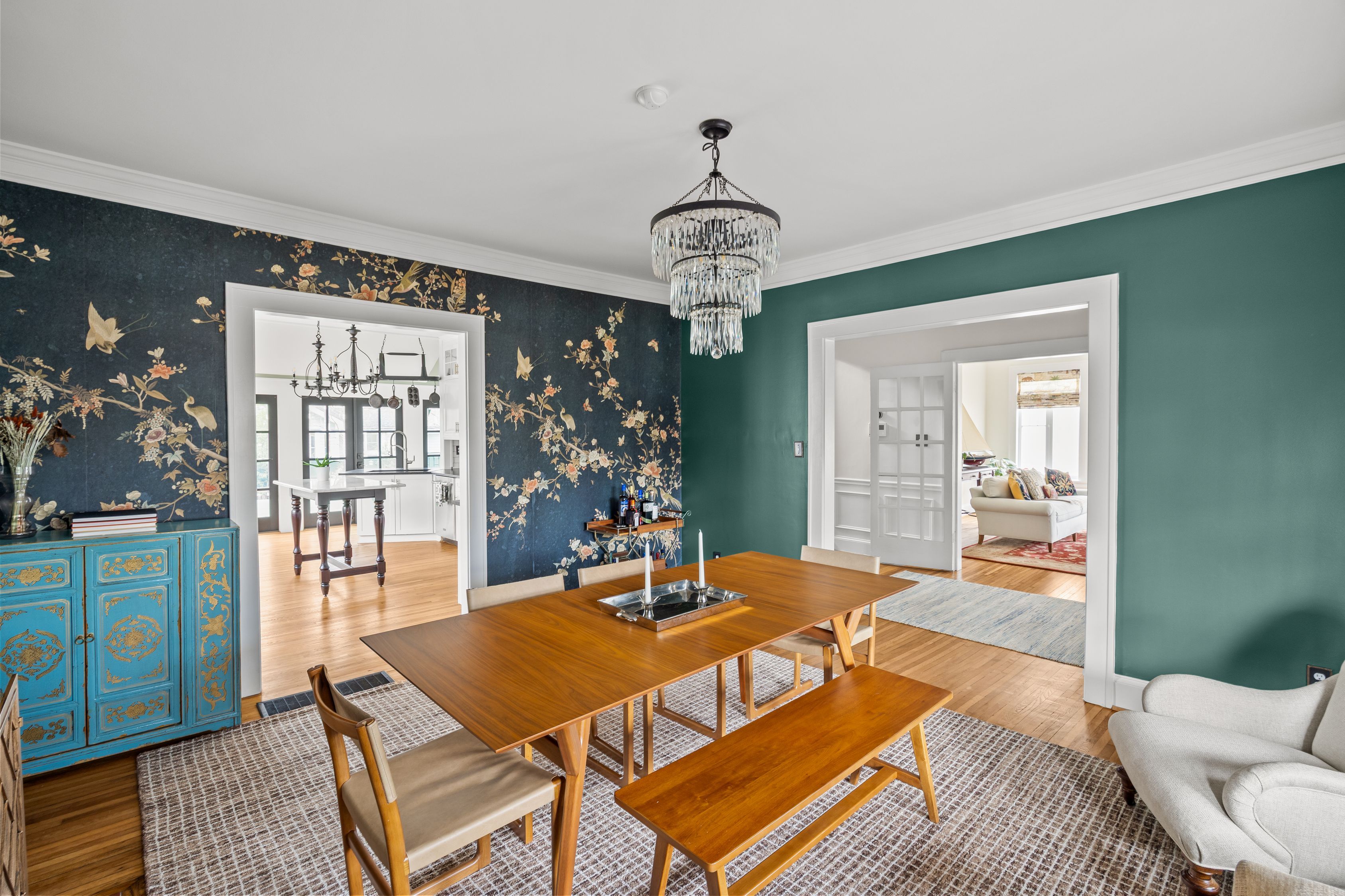Dining room with wooden table and bench, beige chairs, green and floral wallpaper walls, crystal chandelier, blue buffet cabinet, and open doorways to kitchen and living room.