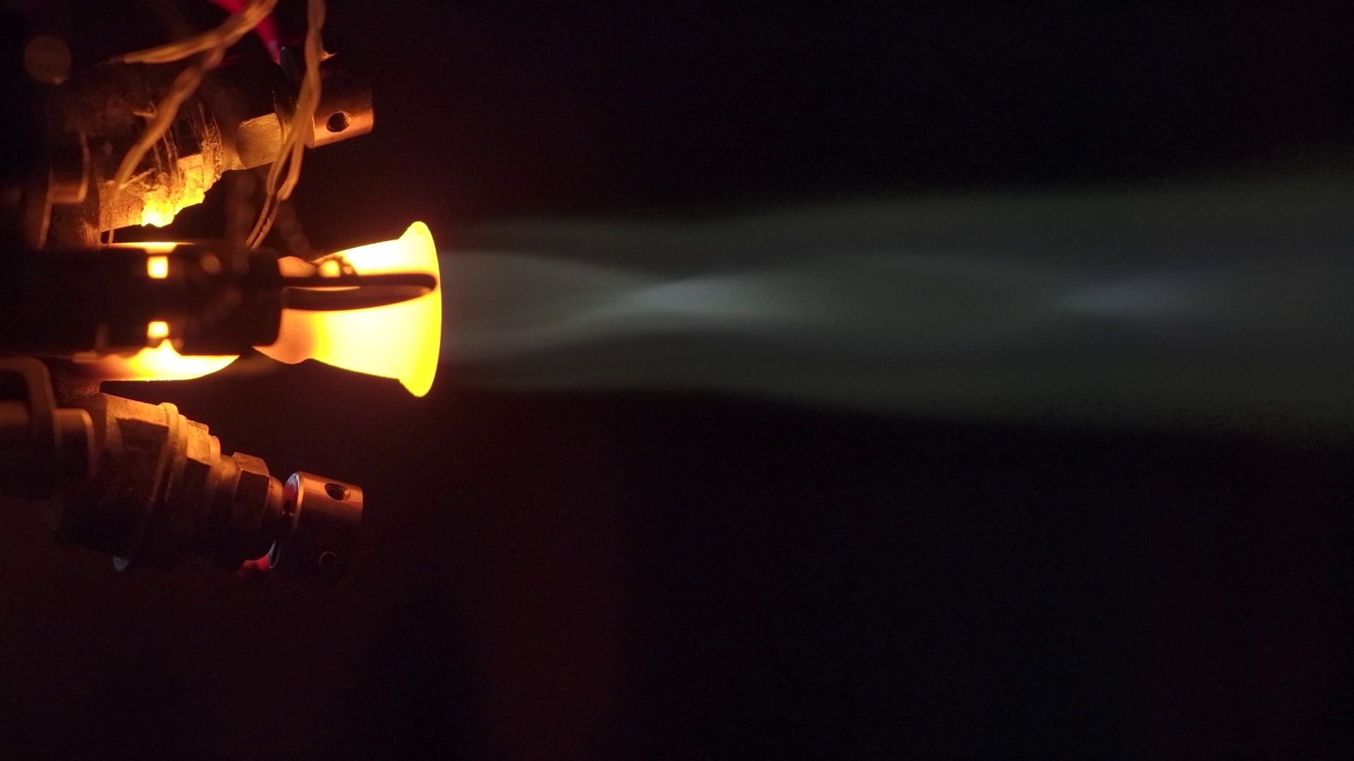 Close-up of a small industrial jet engine nozzle glowing orange and emitting a white exhaust plume against a dark background.