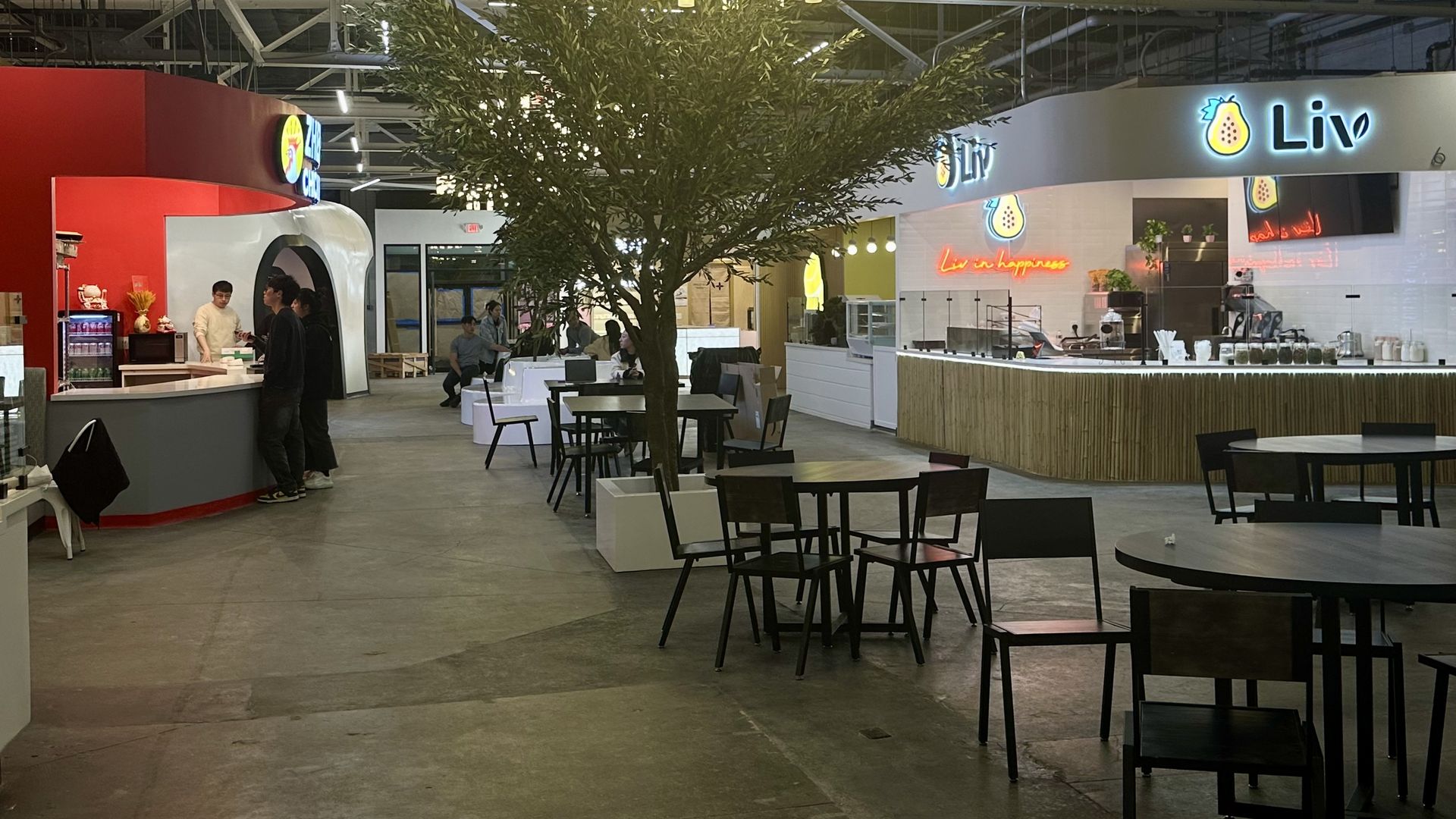 Indoor food hall with a tall tree at the center, a circular chandelier of warm lights overhead, exposed ceiling beams, and a "Liv" cafe on the right; tables and people nearby.