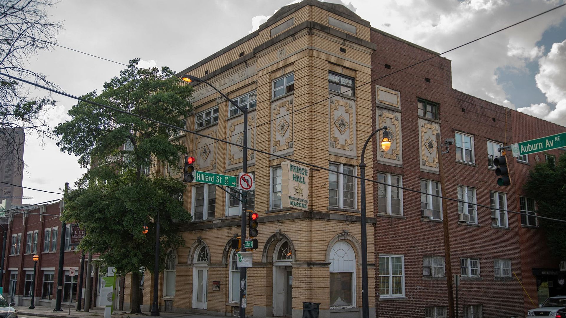 Corner view of a tan brick historic building with arched doorways, a sign reading "Fraternal Hall Free and Accepted Masons," traffic lights, street signs for Hilliard St NE and Auburn Av NE, and some trees.