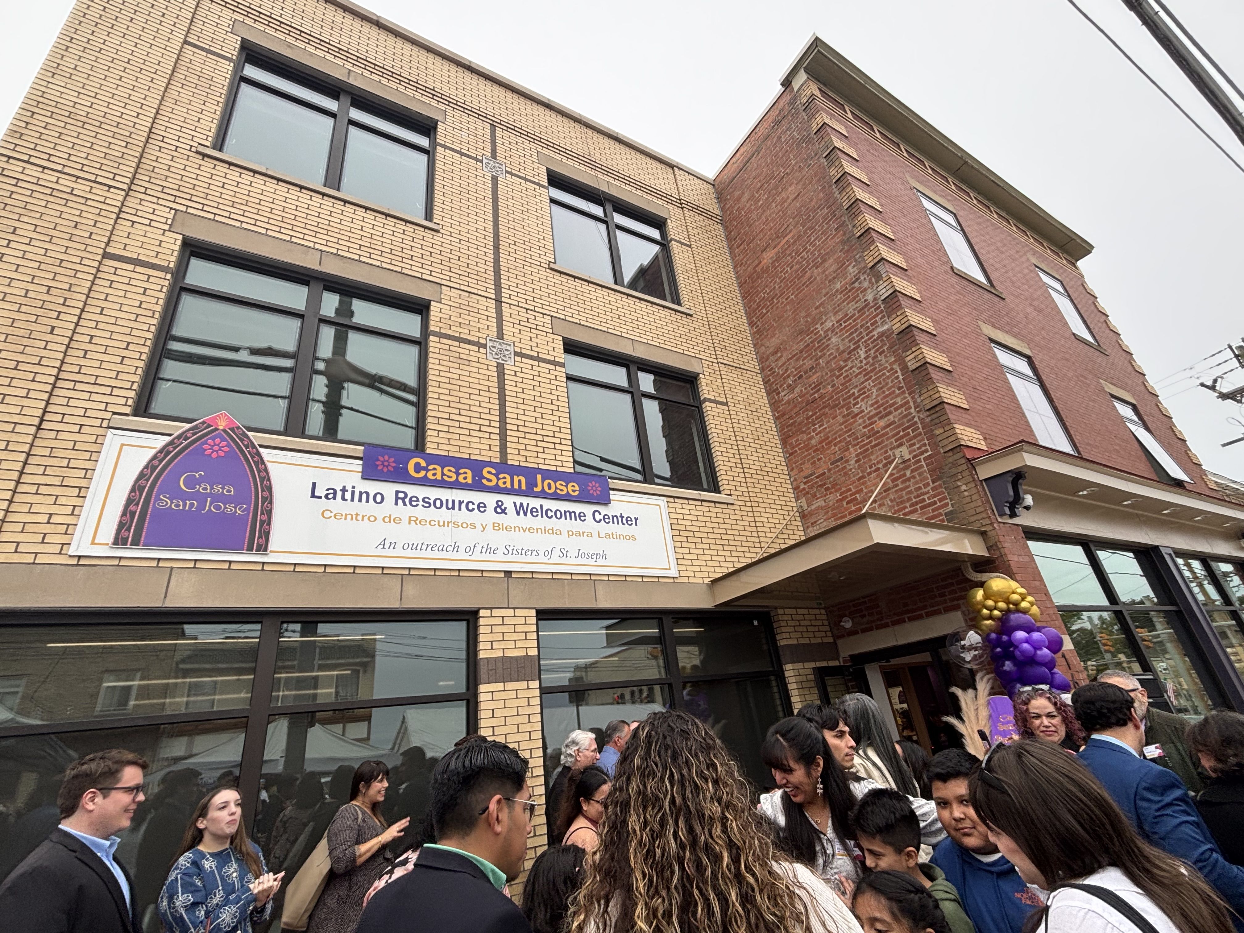 Angled street view of a tan brick building with large windows and a red brick corner. A sign reads Casa San Jose and Latino Resource & Welcome Center; a crowd gathers outside with purple and gold balloons.