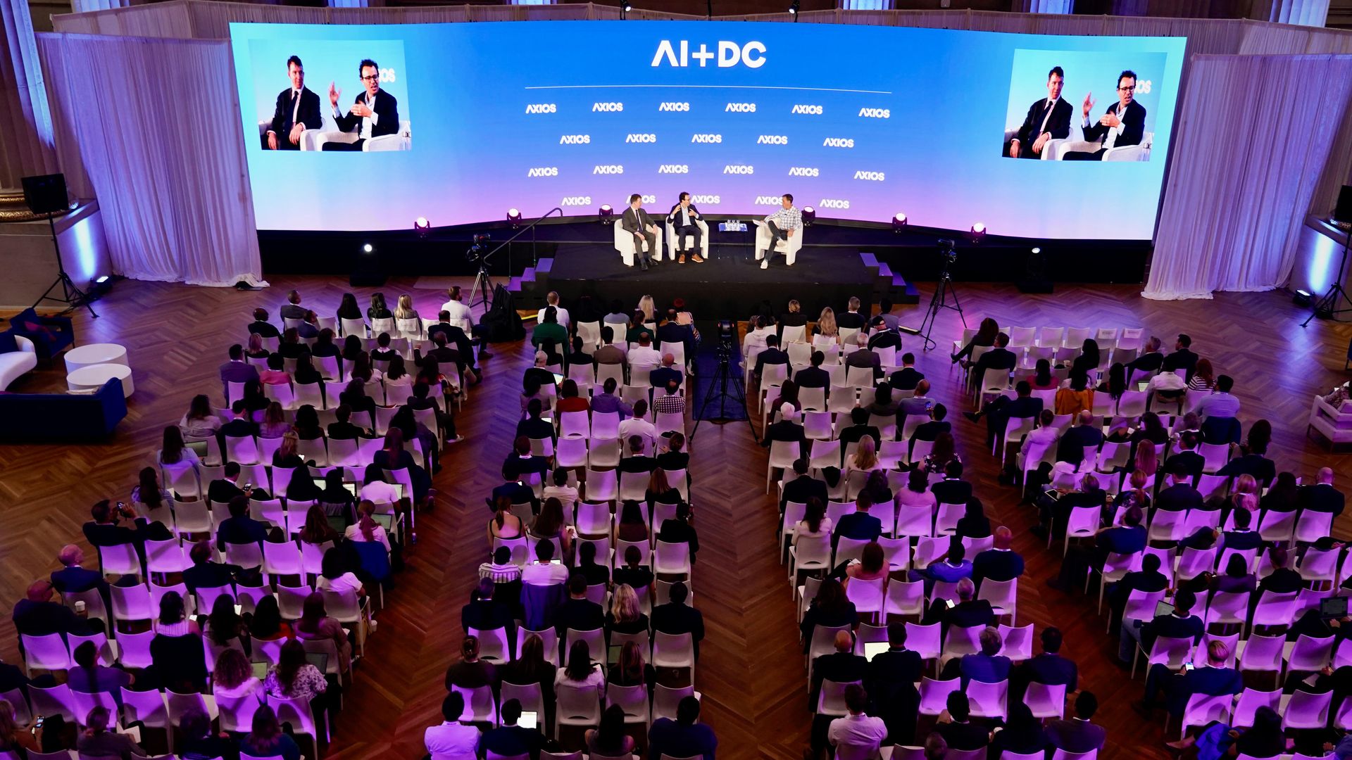 AI+ DC Summit 2025 audience watching three speakers on stage under blue screen,  lit with purple lights.