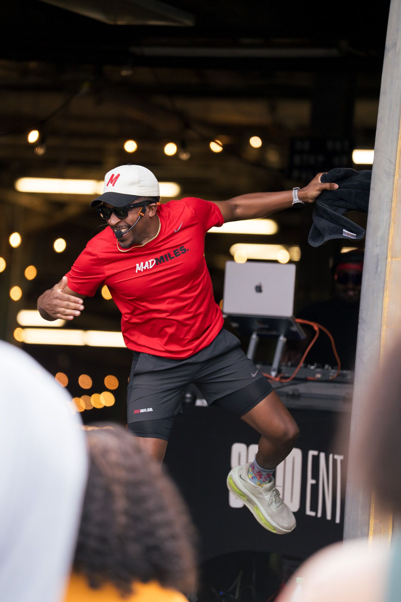 A man in a red MAD MILES shirt, white cap with red M, sunglasses, and a headset mic leans forward, reaching to the right on a stage with warm lights and DJ gear in the background.