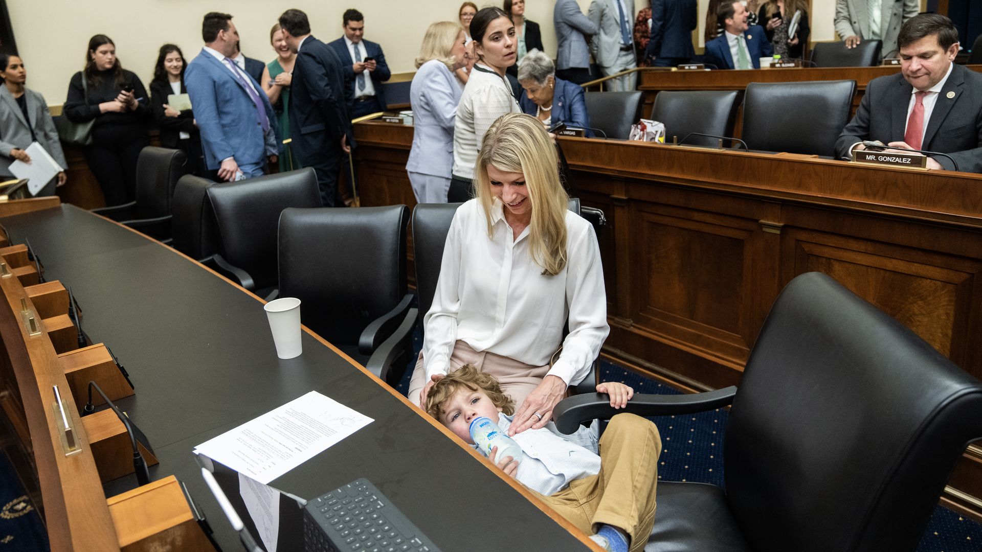 A woman holds her son inside a large room, with several people in business attire in the background. 