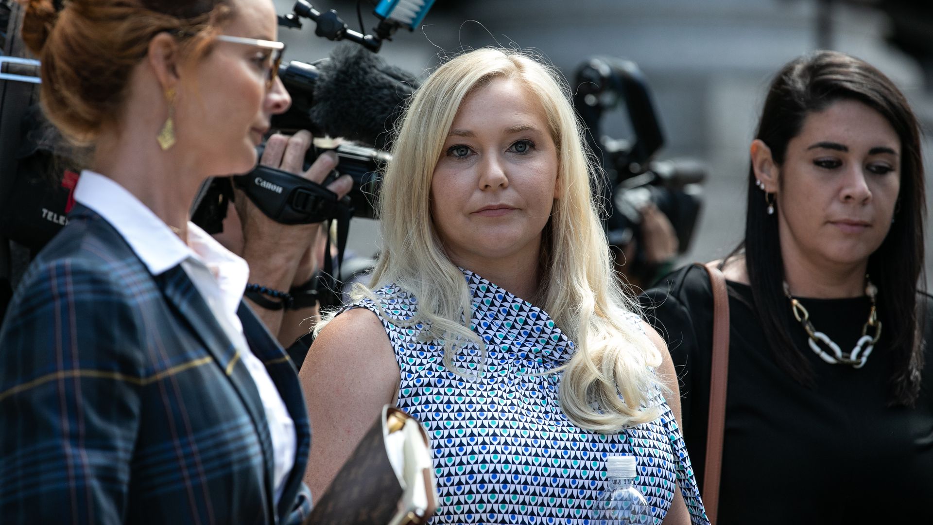 Three women standing outdoors, one with red hair in a plaid jacket, one blonde in a patterned sleeveless top holding a water bottle, and one with dark hair in a black outfit; camera crew filming behind.