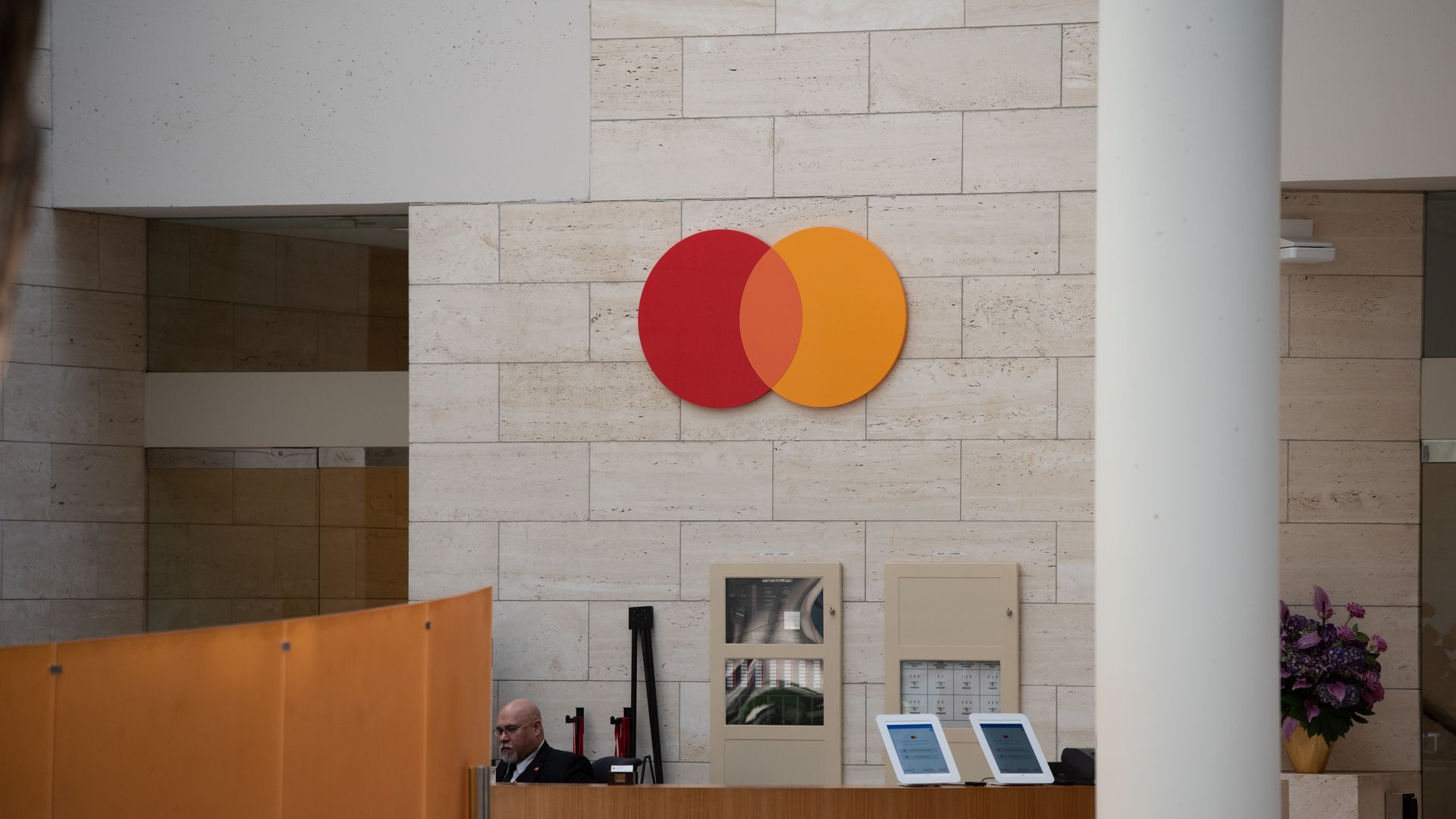 The Mastercard logo, featuring interlocking red and orange circles, mounted on a light-colored stone wall above a wooden reception desk in a corporate lobby. 
