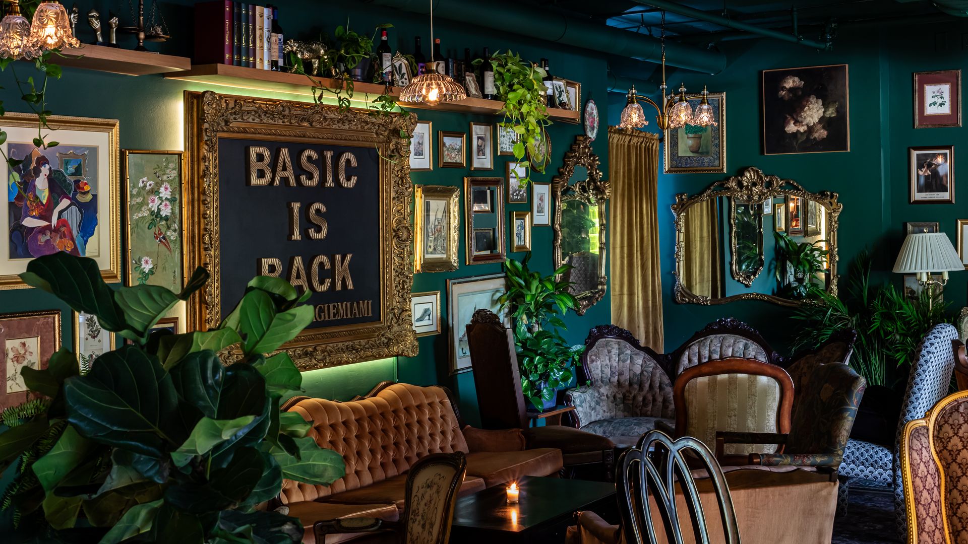 Teal-walled vintage lounge with ornate gold picture frames and a collage of art prints. Crystal chandeliers hang above shelves of bottles and trailing plants, with tufted sofas and mirrors.