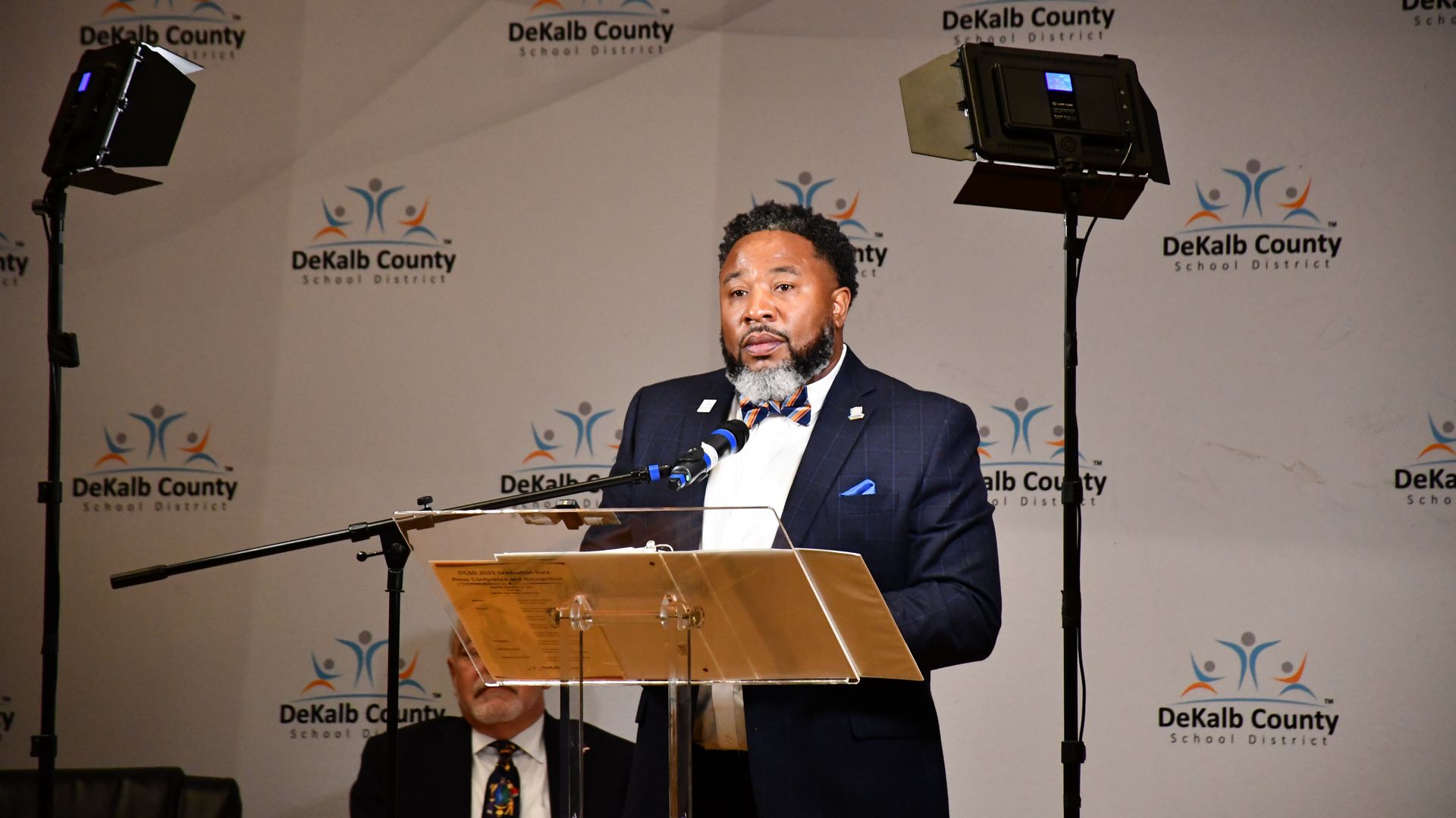 A man in a dark suit and bow tie stands at a clear podium speaking into a microphone against a DeKalb County School District backdrop with studio lights on either side.