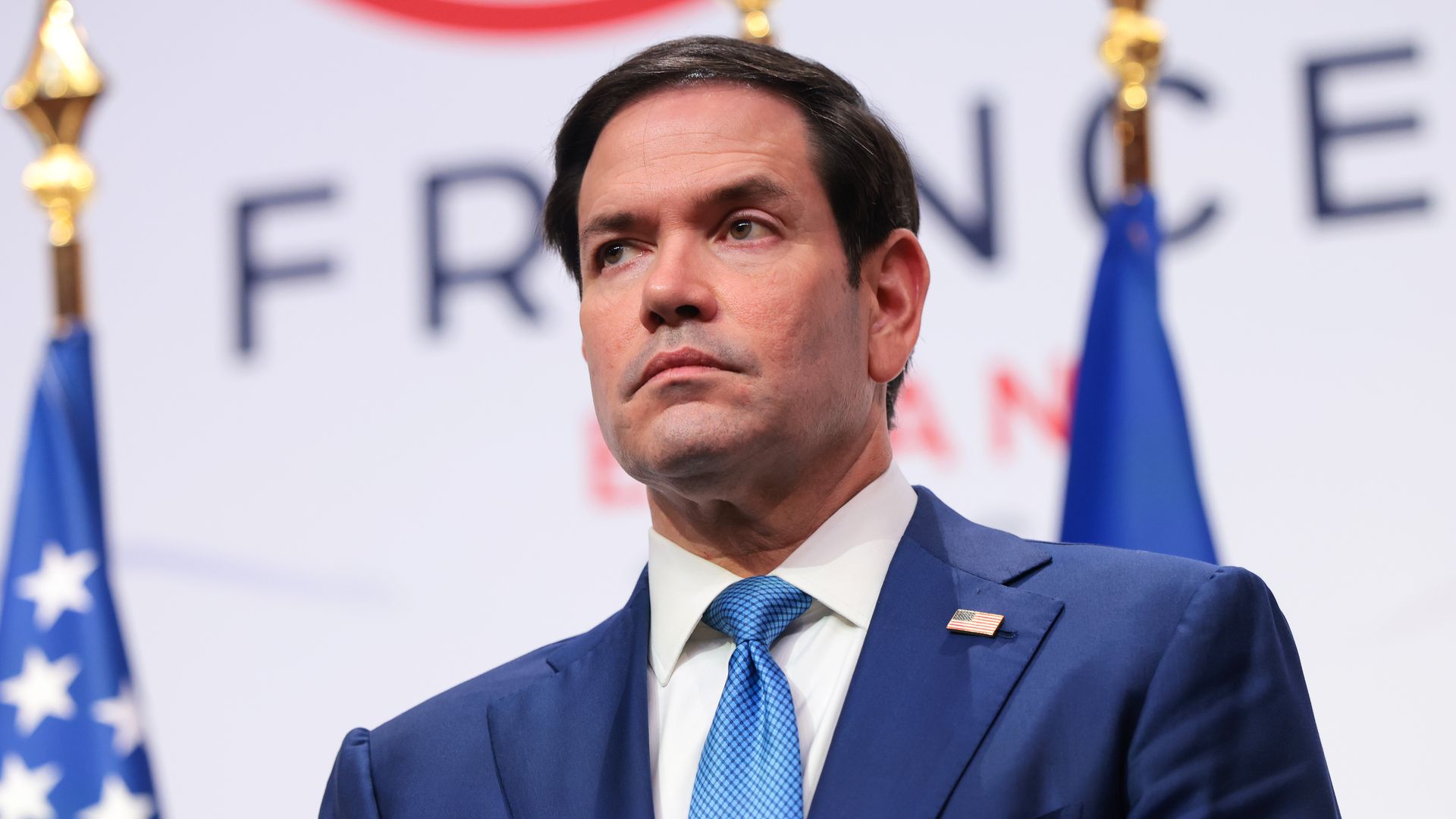 Man in a blue suit and light blue tie with an American flag pin, at a podium, looking to the left, against a backdrop of blurred flags.