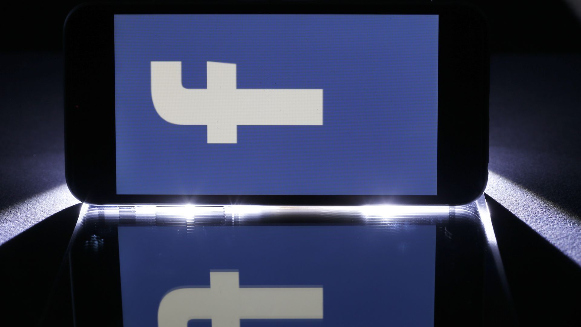 Photo of phone on its side on reflective table, showing a giant Facebook symbol on its screen