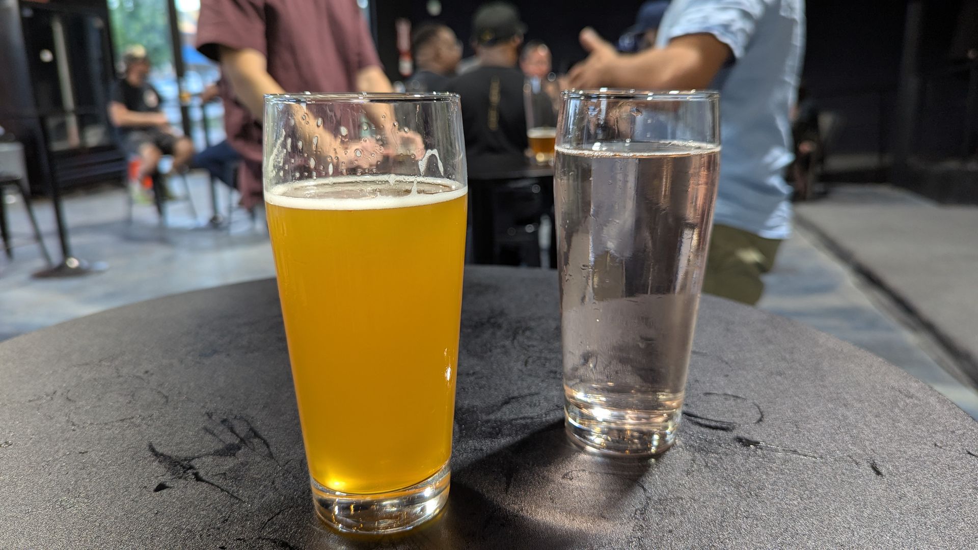 A glass of beer and a glass of selzer on a bar table with people at tables in the background.