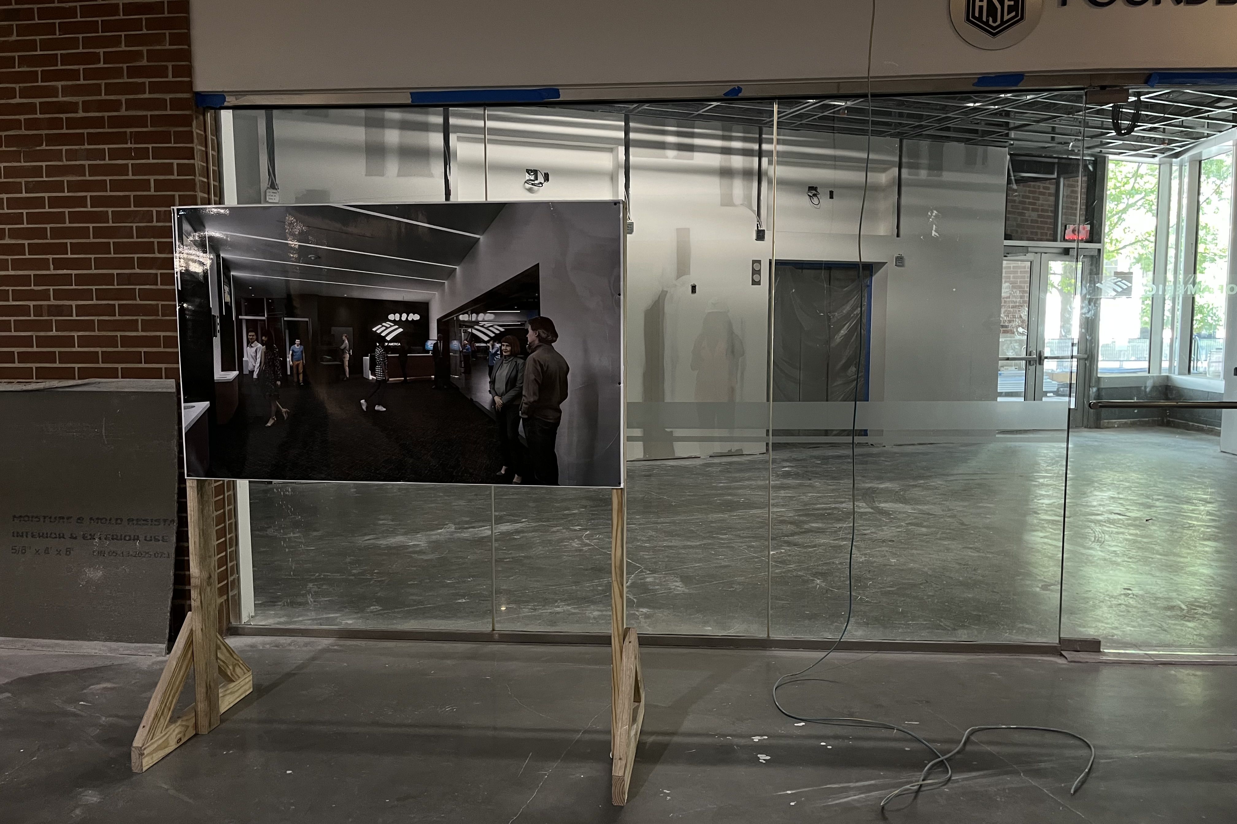 Construction site of an unfinished commercial space with glass walls and concrete floor, a wooden easel holding a rendering of the completed interior with people inside, exposed wiring.