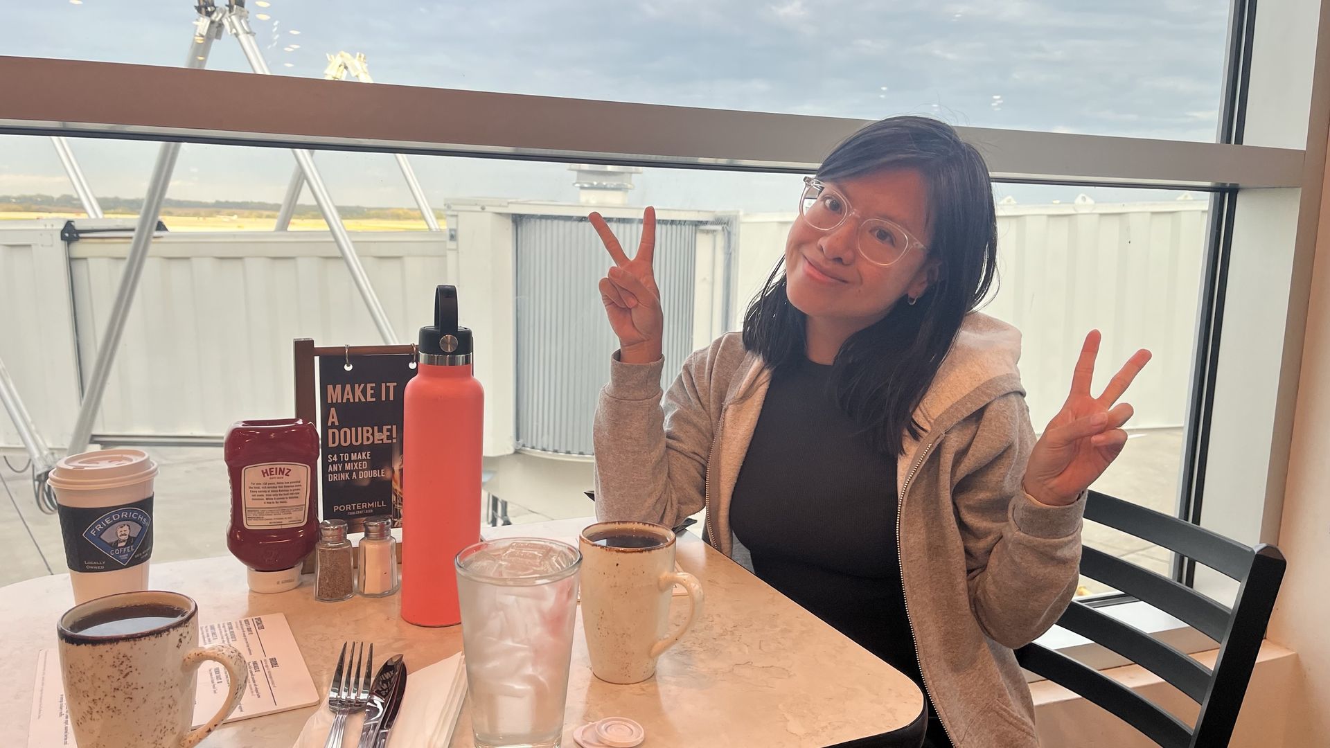 Linh with glasses and black hair wearing gray hoodie and black shirt, seated at table in Portermill at the Des Moines Airport near window, making peace signs with both hands. Table holds beverages, silverware, and condiments.
