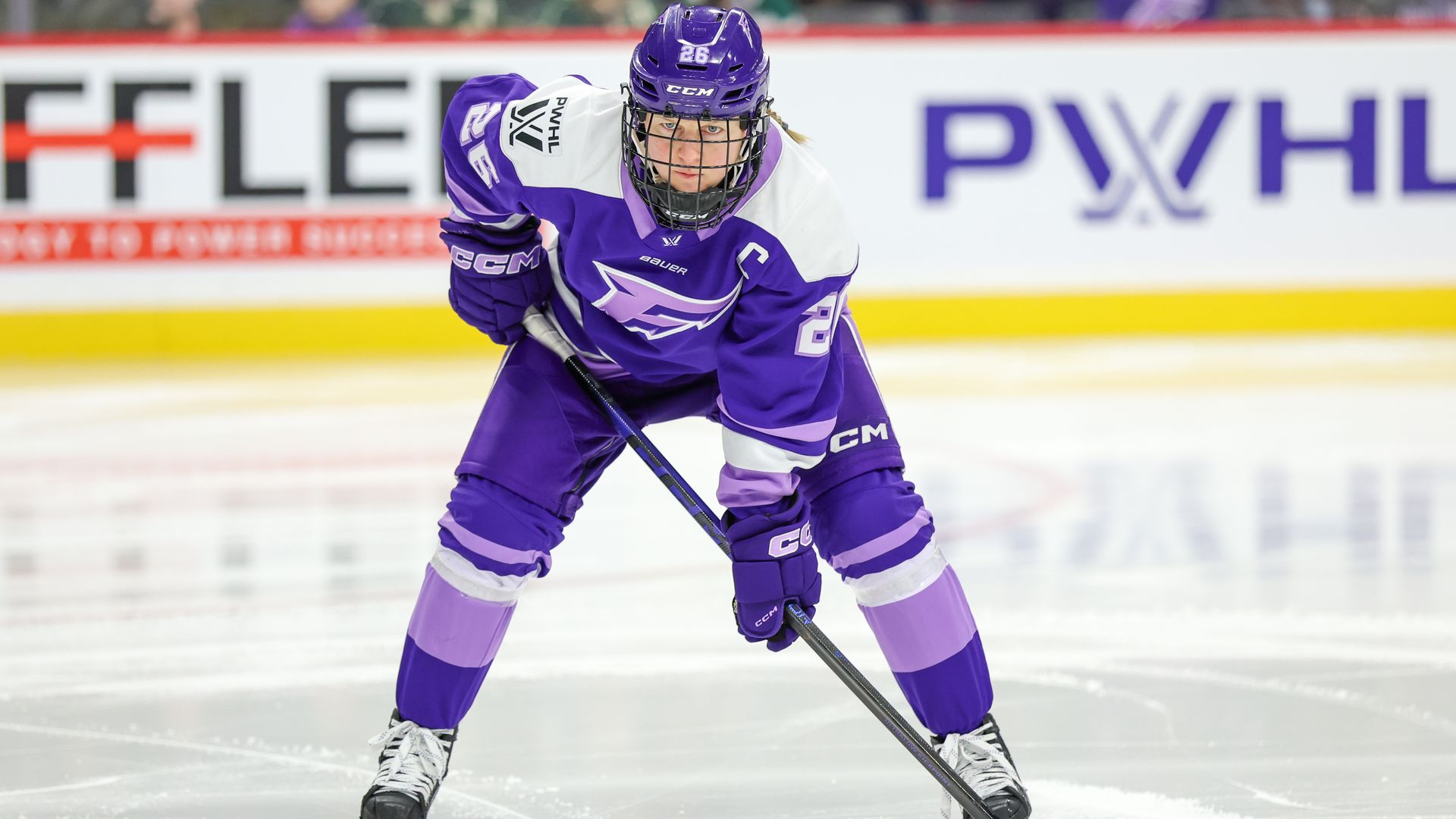 Ice hockey player in a purple uniform (number 26) with gloves and helmet, skating and handling a stick toward the puck on the rink, with a crowd and sponsor boards in the background.