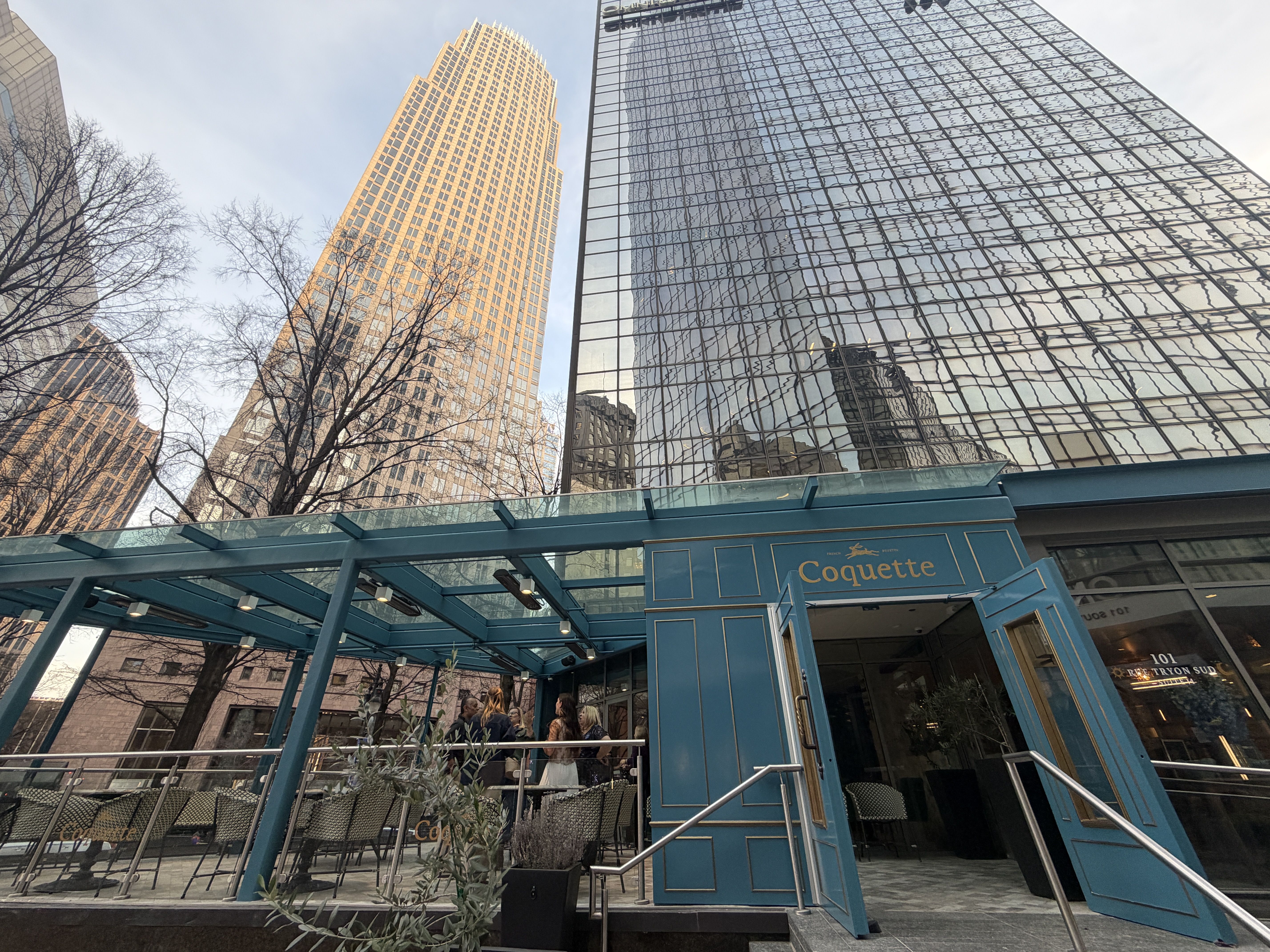 Blue storefront with gold Coquette sign beneath a glass canopy, outdoor seating along a railing. Bare trees frame tall glass skyscrapers reflecting in the glass facade.