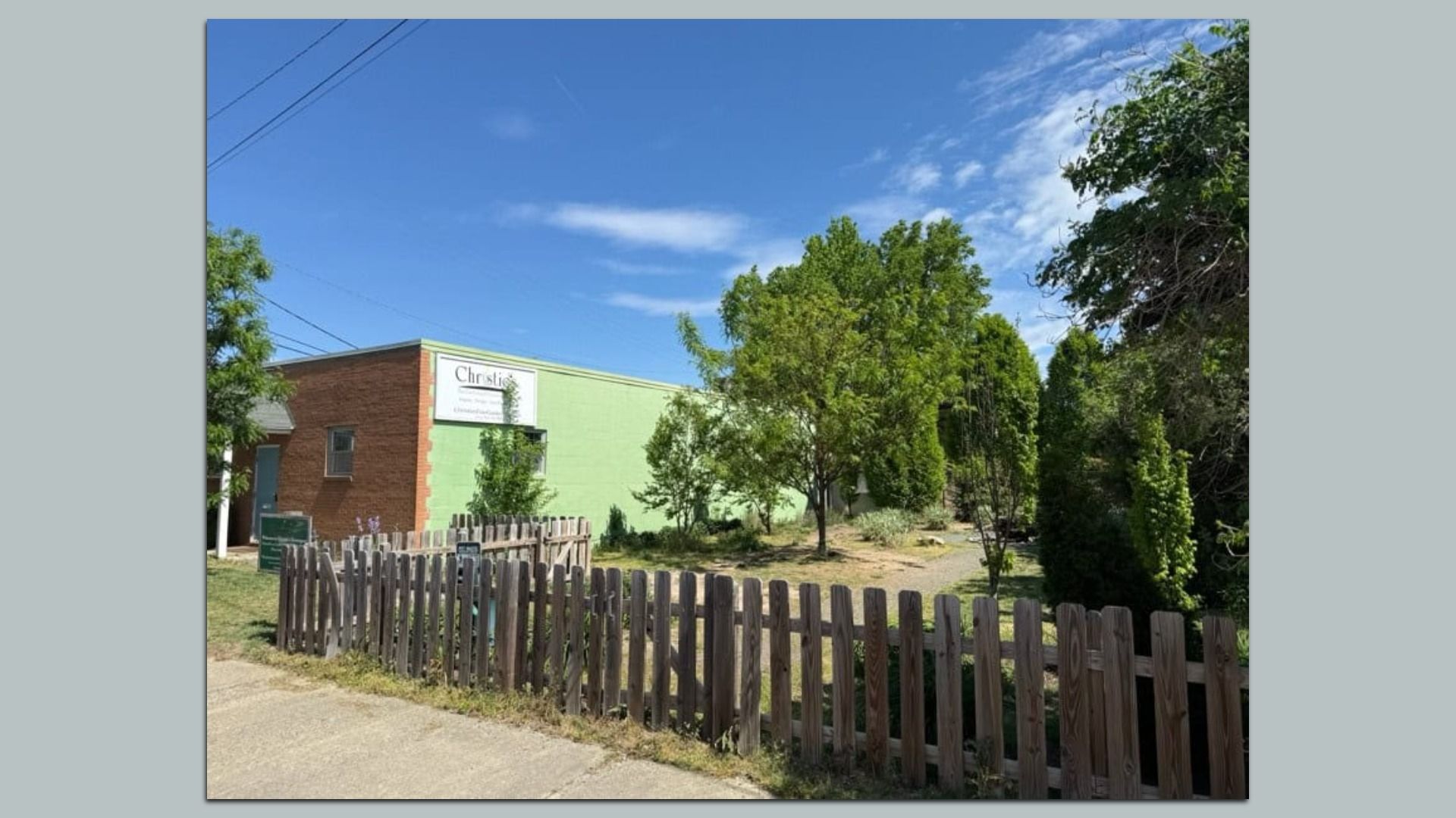 Exterior view of a small commercial building with a pale green wall and brick accent, a wooden picket fence in front, several green trees, a blue sky with wispy clouds, and overhead power lines.