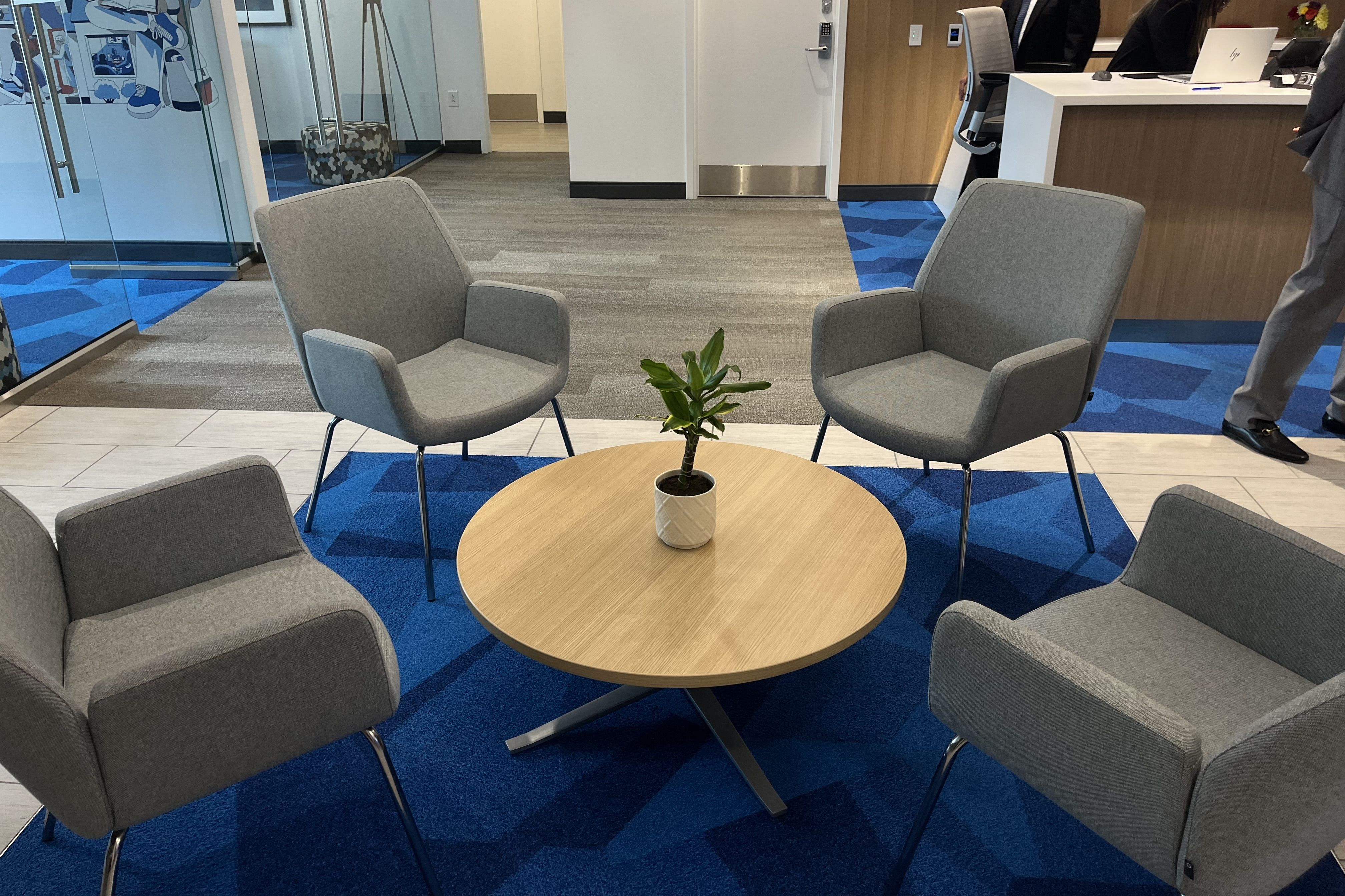 Four gray armchairs arranged around a round wooden table with a small potted plant on top, set on a blue carpet in a modern office lobby with glass walls and people at a reception desk.