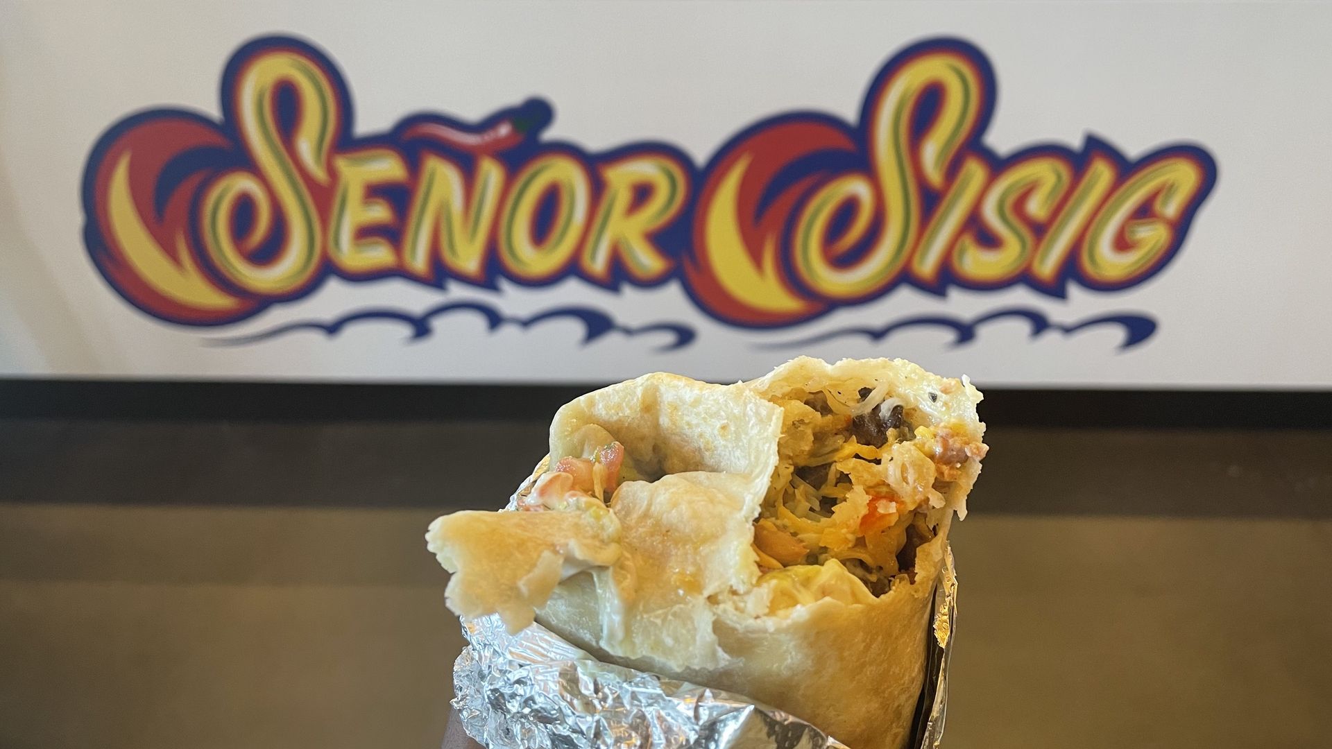 a burrito with a bite taken out of it in front of a sign that says "senor sisig"
