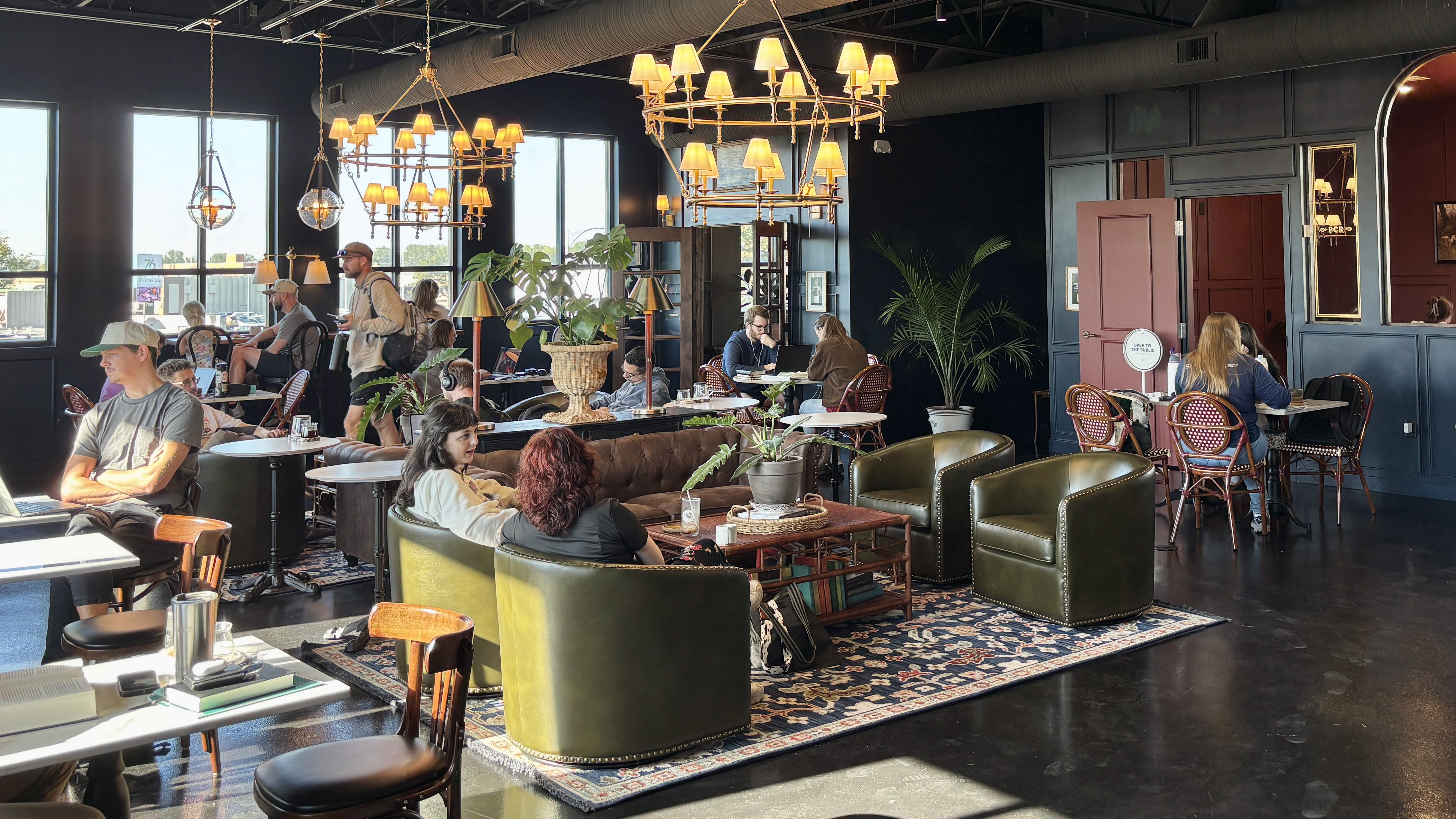Stylish cafe interior with dark blue walls, tall windows, and large chandeliers. Bronze and wicker seating surrounds tables where people work on laptops or chat, with potted plants nearby.