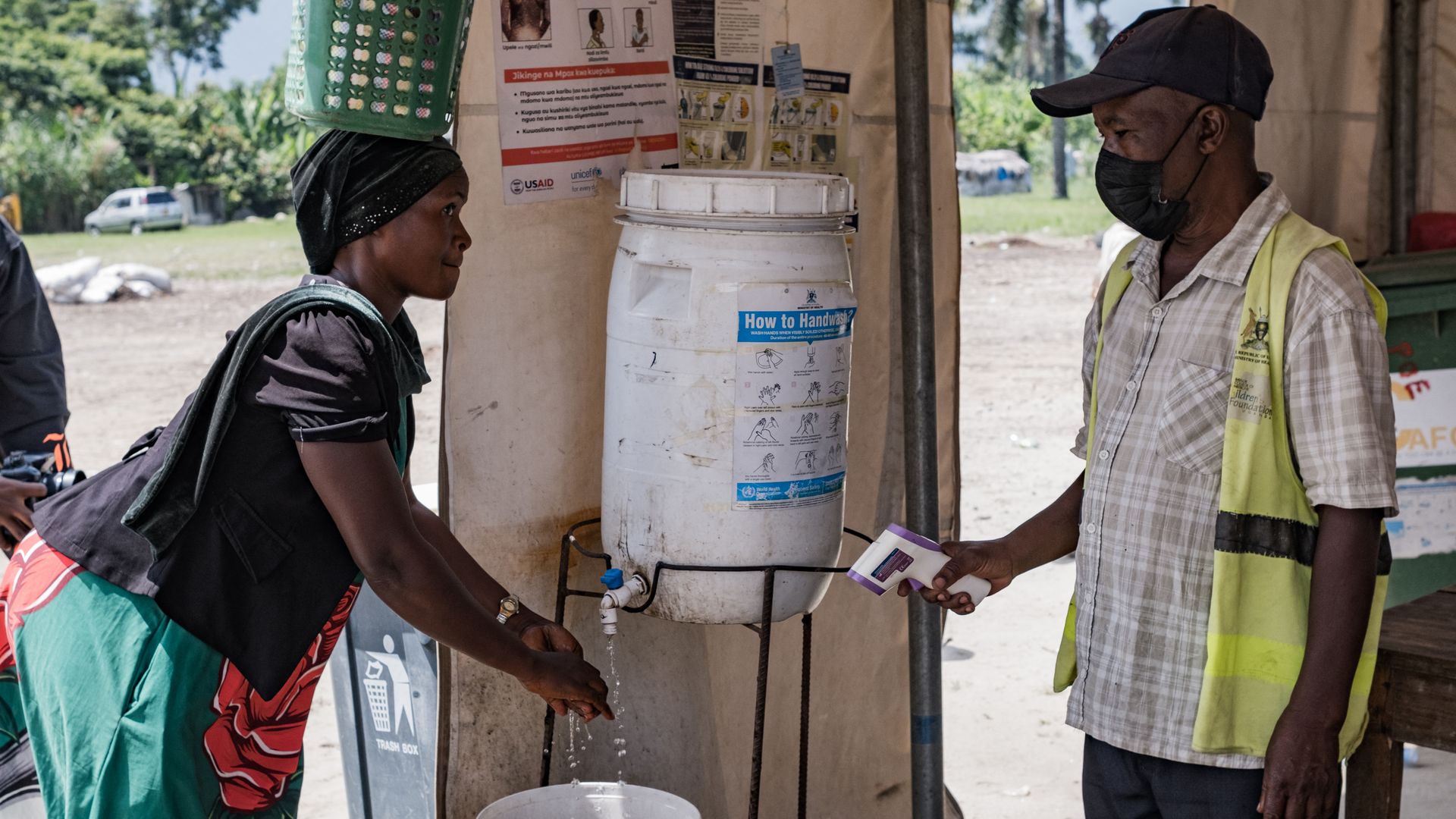 Woman in a bright dress balances a green basket on her head while washing hands at a white handwashing station; a man in a cap and mask wearing a yellow vest stands nearby with posters and a blue-labeled bucket.