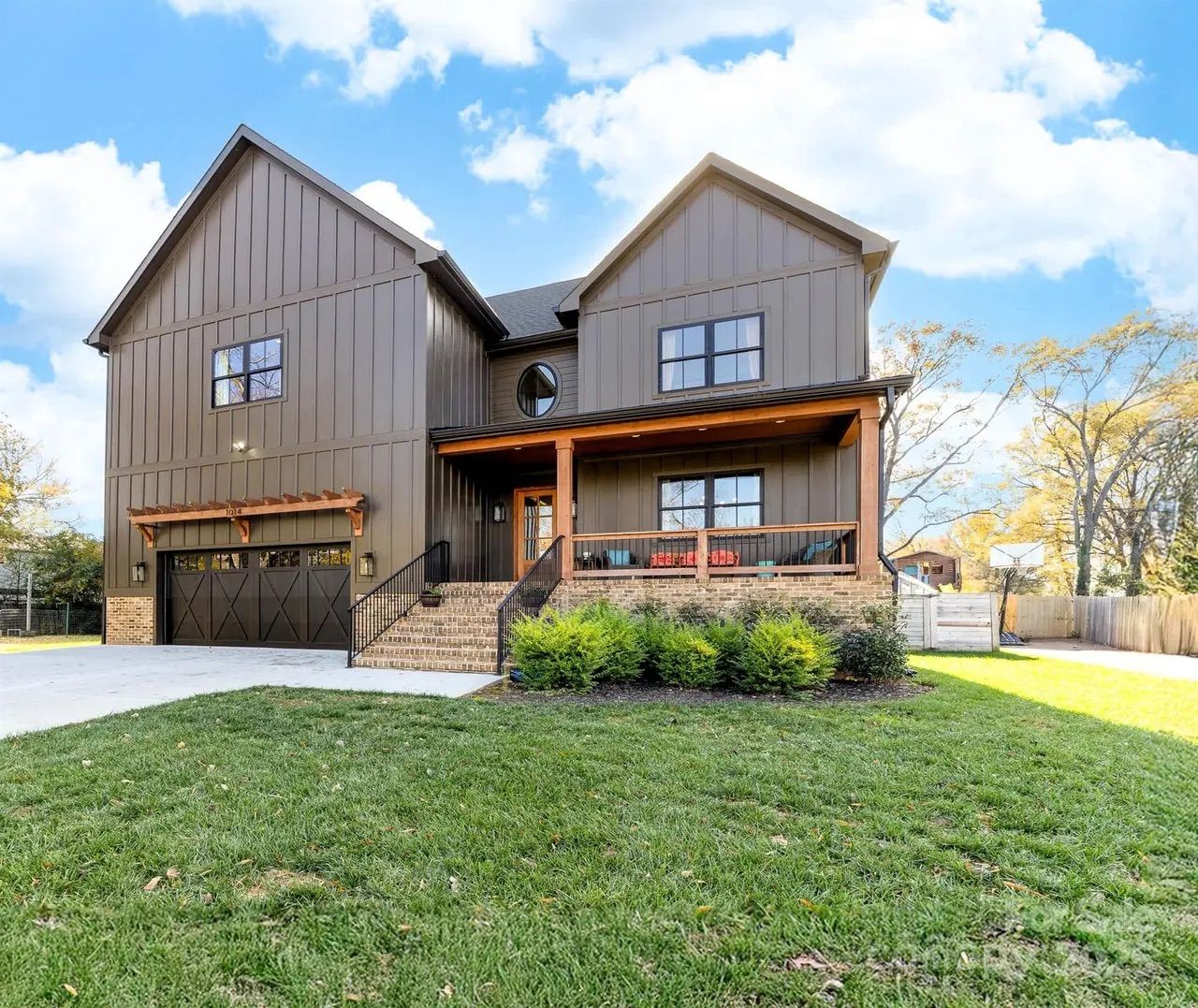 Modern two-story brown house with brick foundation, large garage, front porch with red and blue cushions, green lawn, and blue sky with some clouds.