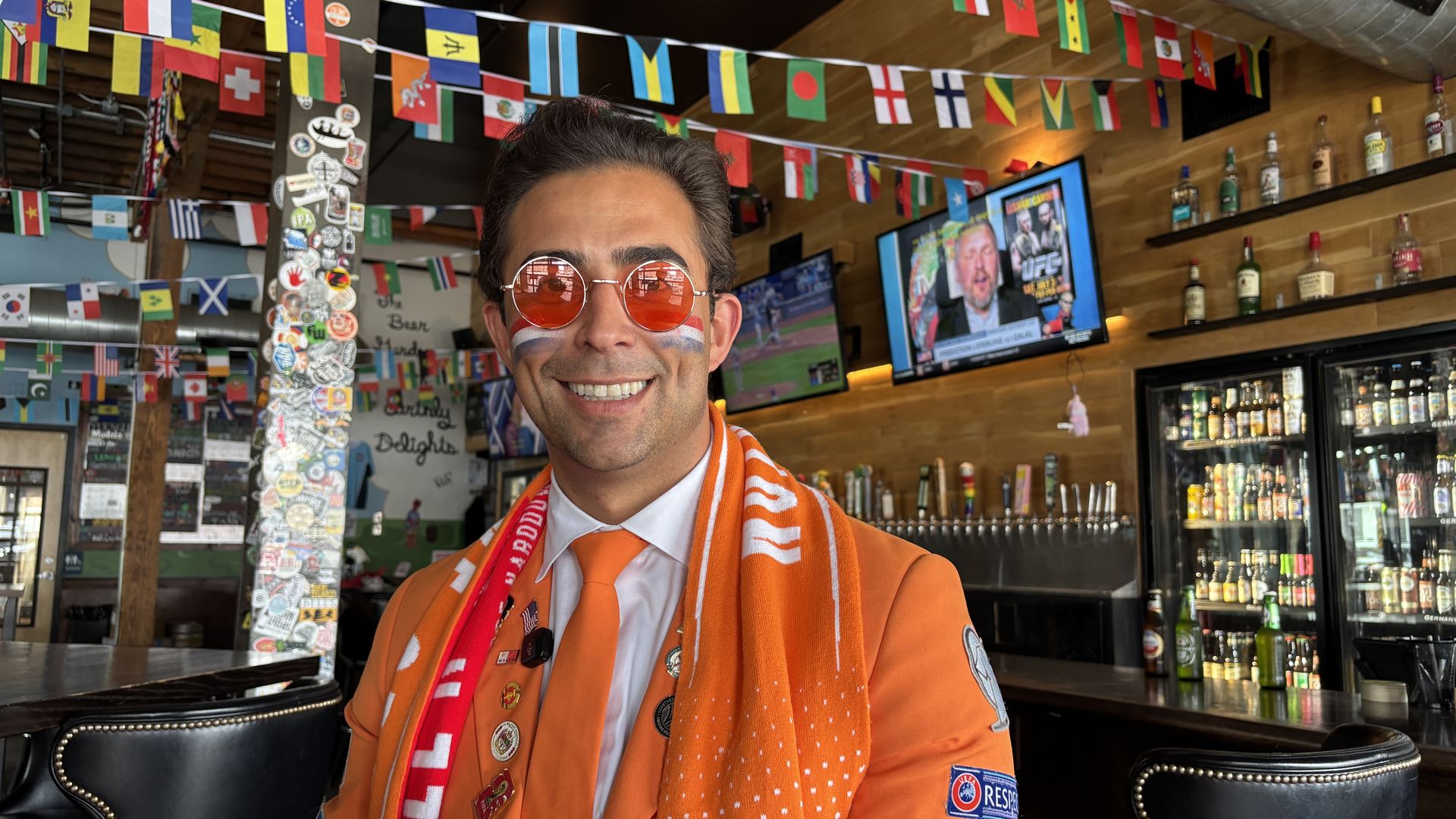 Smiling man in an orange suit with an orange scarf and round red sunglasses stands in a bar, surrounded by hanging international flags, with liquor shelves and a TV screen in the background.