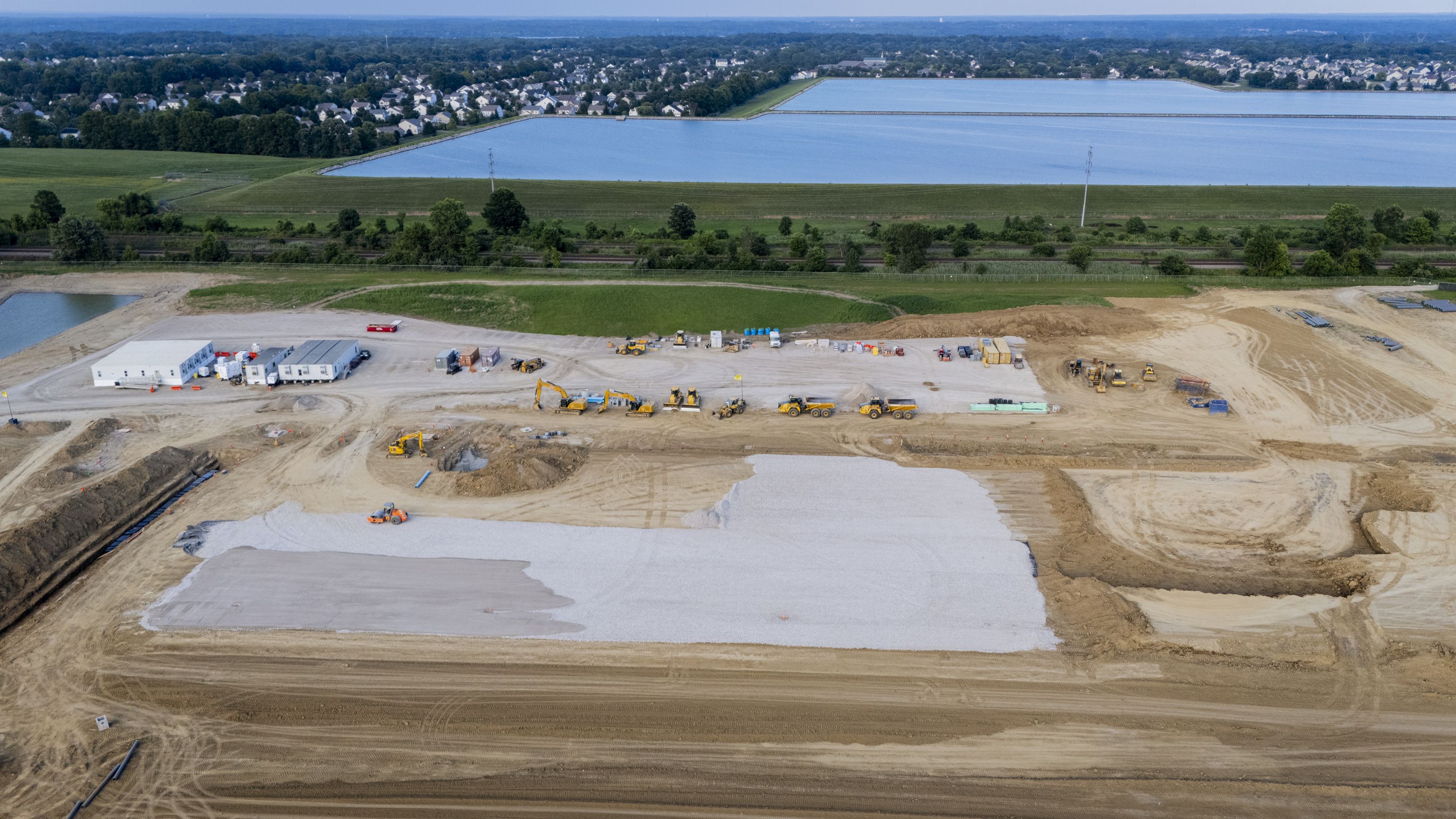 A large construction site with yellow machinery, dirt, and gravel areas, temporary white buildings, and a residential neighborhood with water bodies in the background under a clear blue sky.