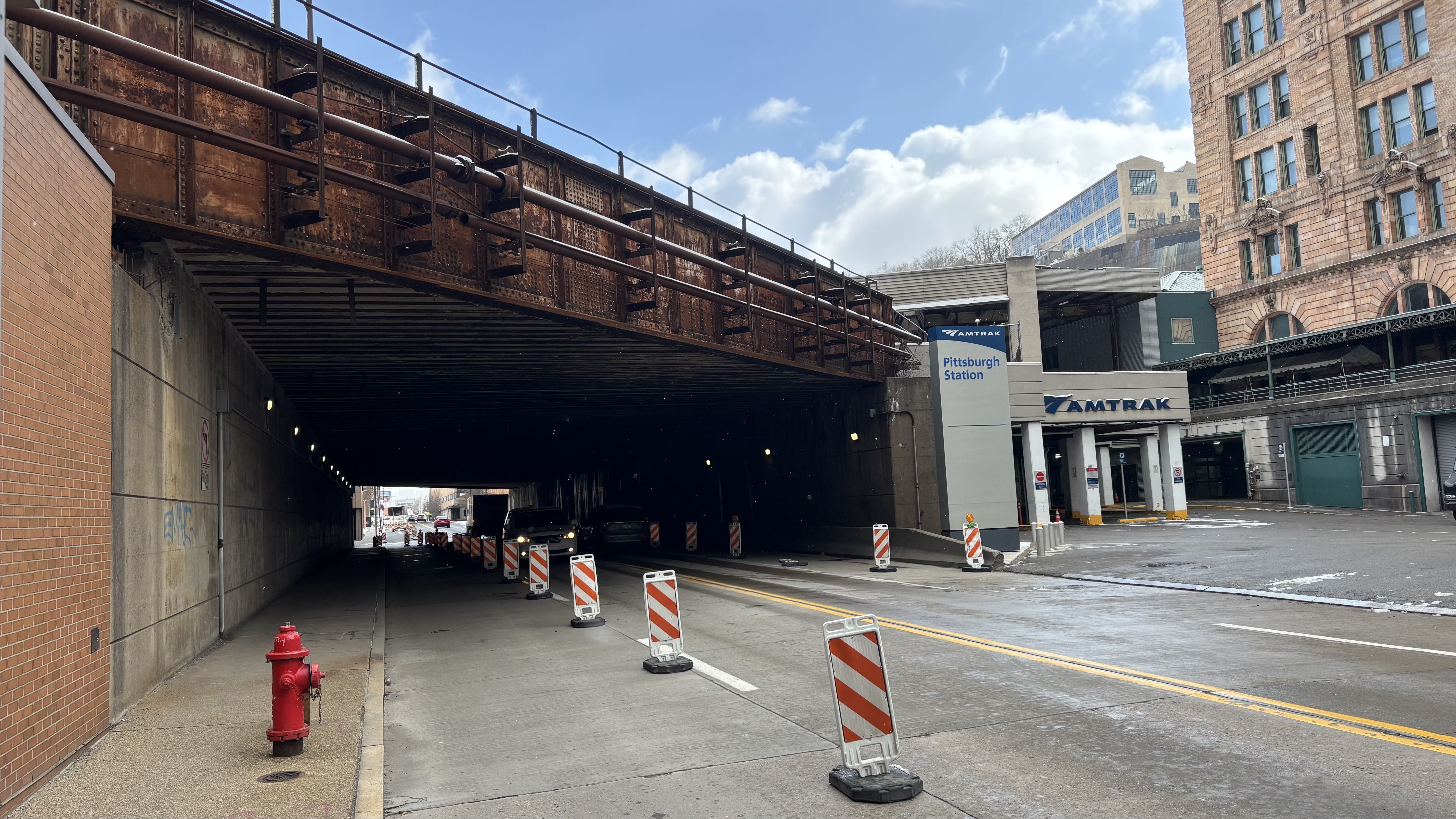 Under a rusted iron railroad bridge over a city street, with orange-and-white barriers. A brick building on the right and the Amtrak Pittsburgh Station entrance ahead; a red fire hydrant on the left.