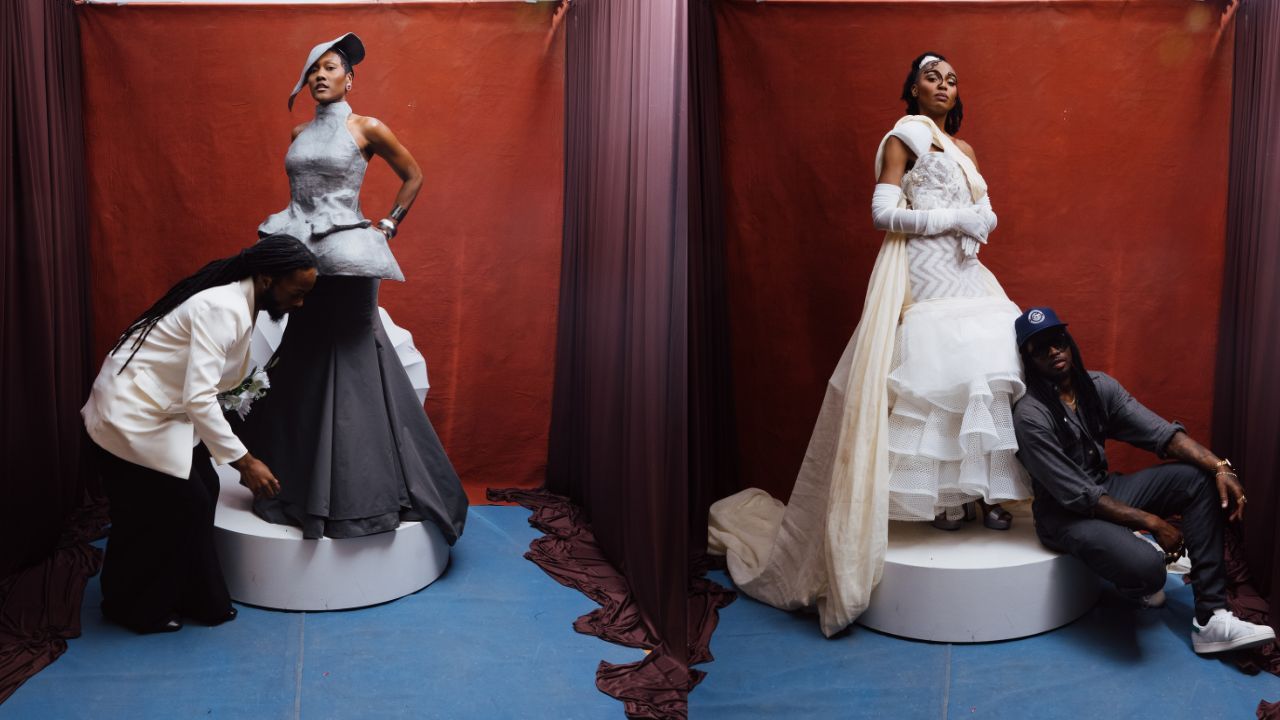 Two models on circular platforms before red drapes. Left: woman in gray peplum gown and hat while a man adjusts her dress. Right: woman in white layered wedding gown with gloves; another man sits beside.