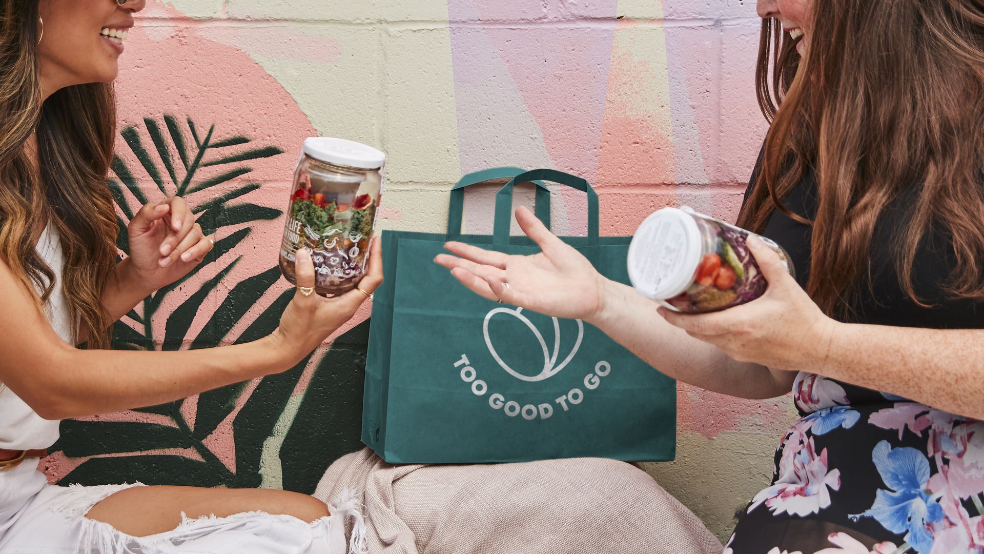 Two women hold jars stuffed with veggies in front of a Too Good To Go-branded recyclable bag. 