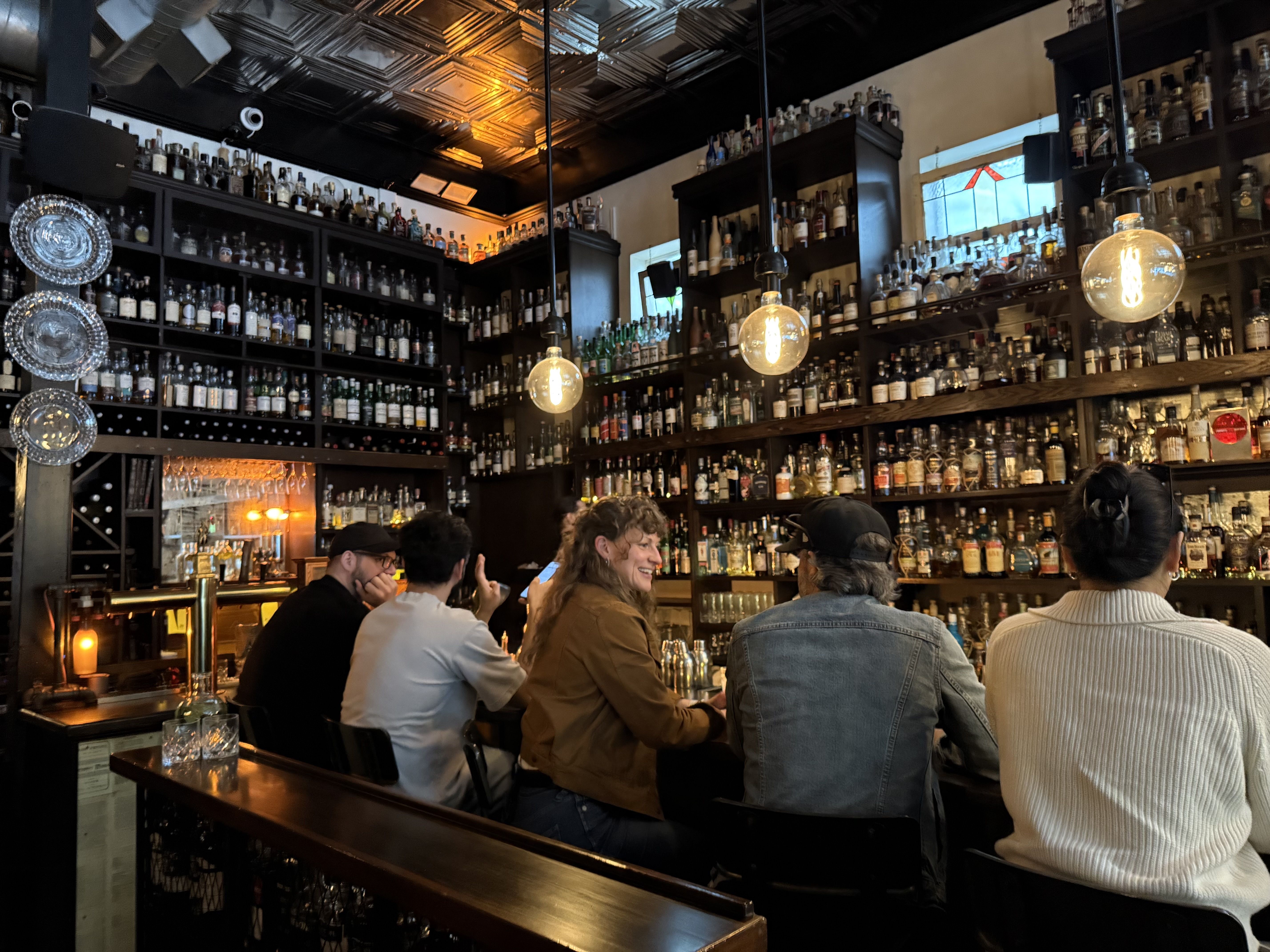 Dimly lit bar interior with dark wood shelves full of bottles, warm hanging bulbs, and patrons seated at the counter chatting and smiling.