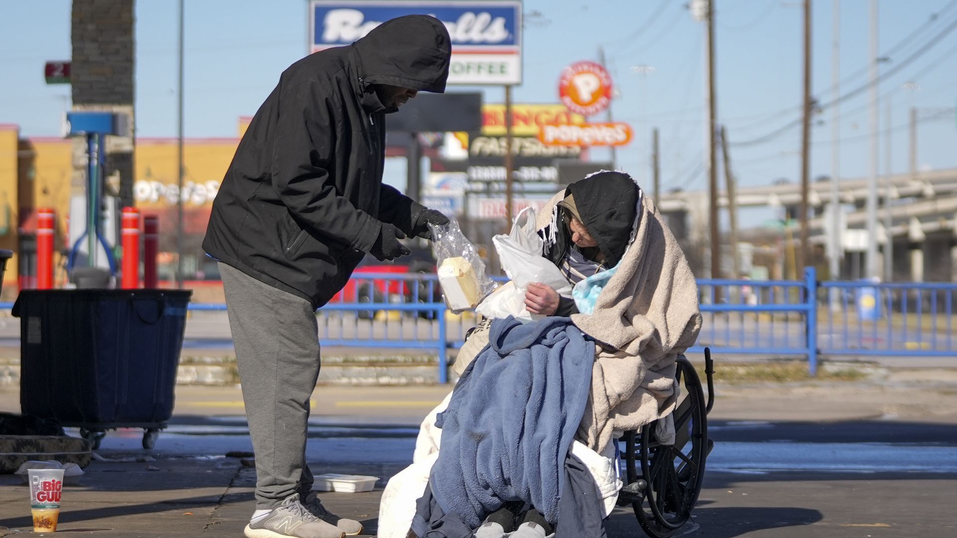 A hooded person leans toward a person in a wheelchair, handing over items from a plastic bag; both wrapped in blankets on a city sidewalk, with signs, power lines, and a Big Gulp cup nearby.