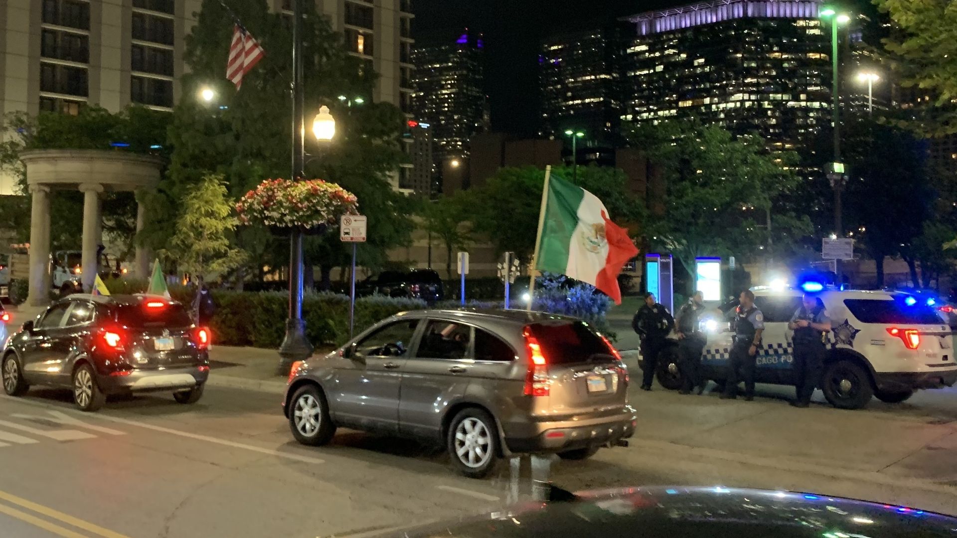 Night city street scene with two cars, one flying a large Mexican flag. Several police officers stand near a police car with flashing blue lights. Buildings and trees in background.