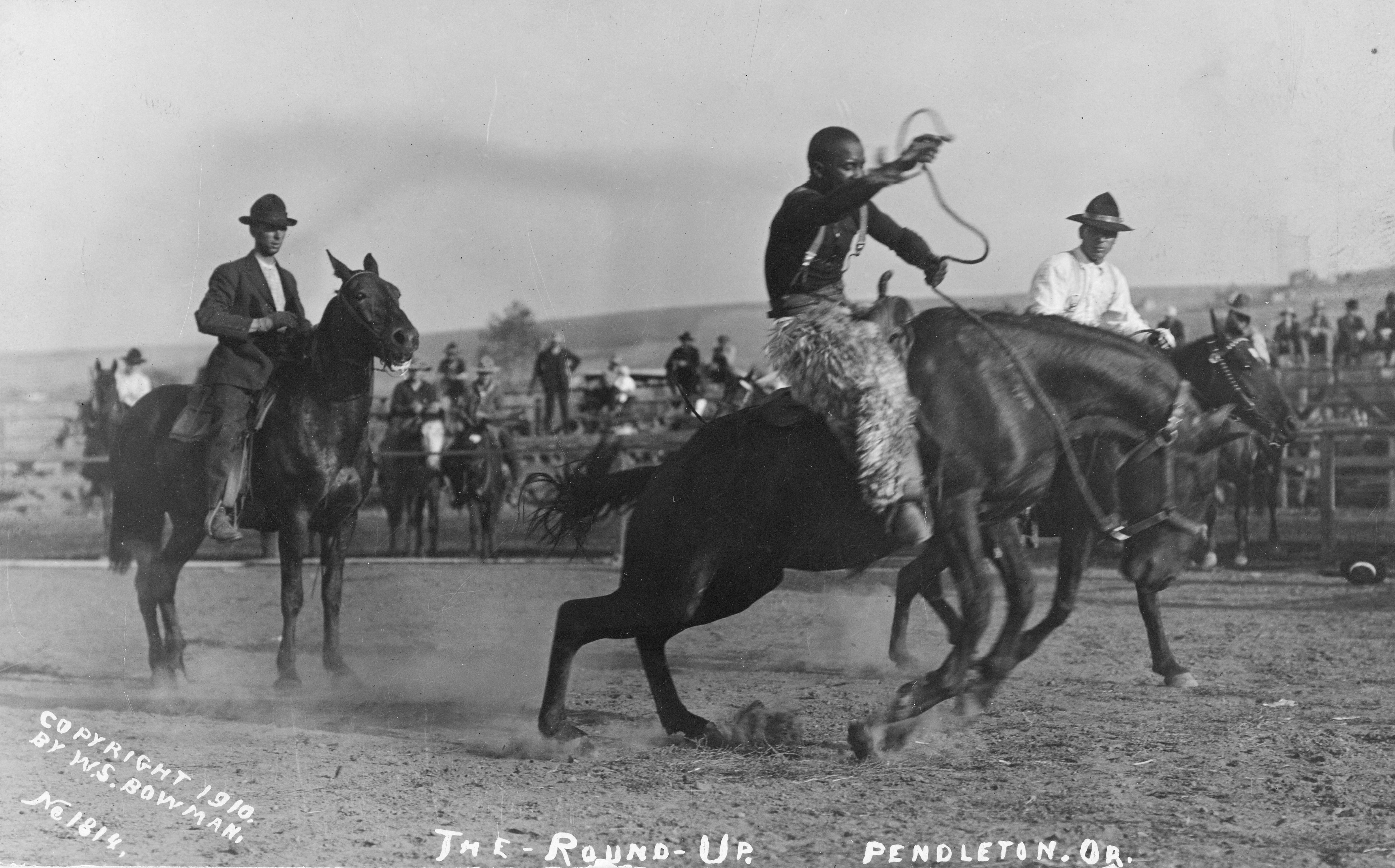 Photo of African-American cowboys riding horses in a rodeo area, with spectators and other participants in the background.