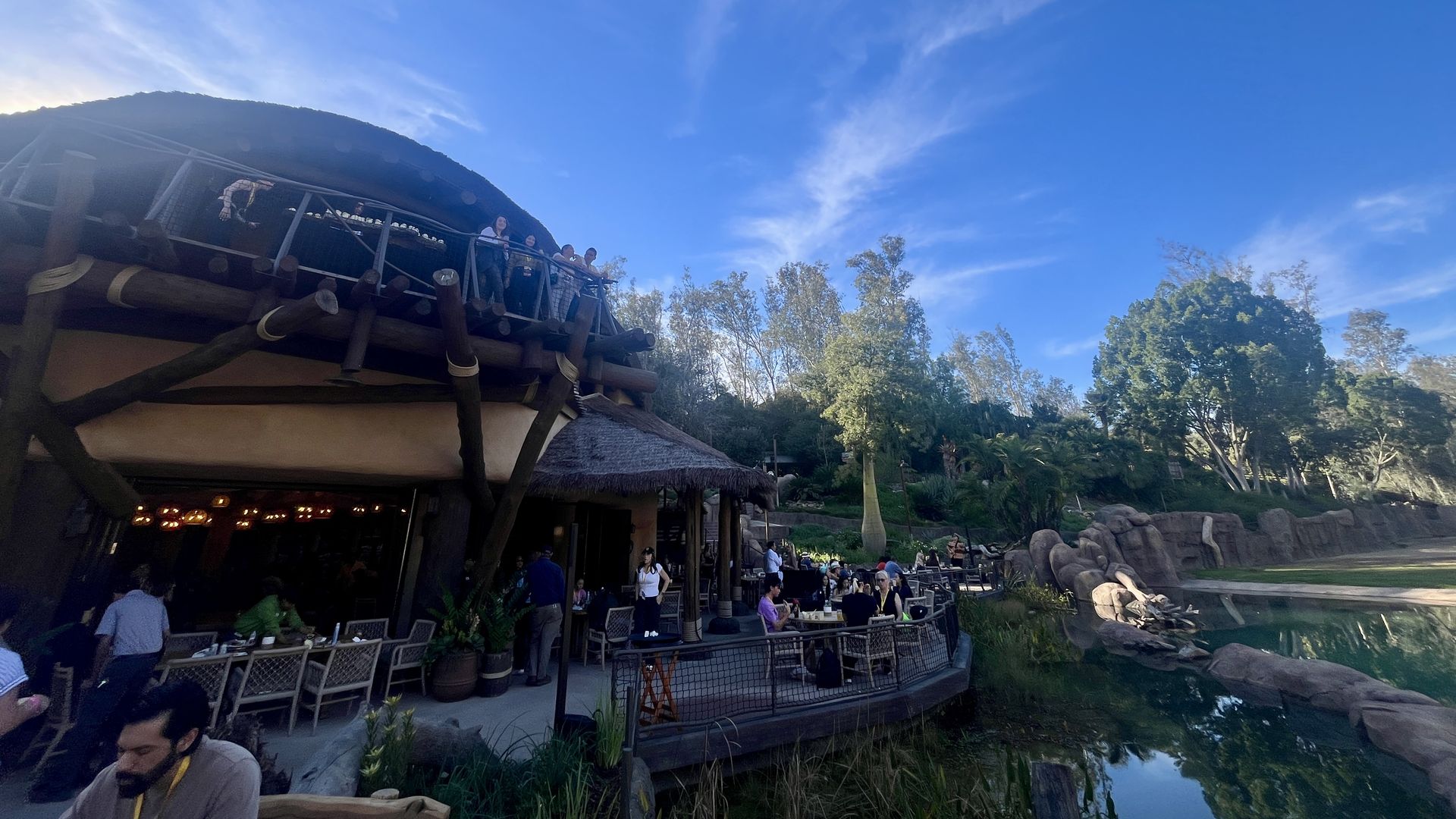 Outdoor dining area with people seated and standing near a rustic wooden building with a thatched roof, surrounded by trees and a calm pond under a bright blue sky with wispy clouds.