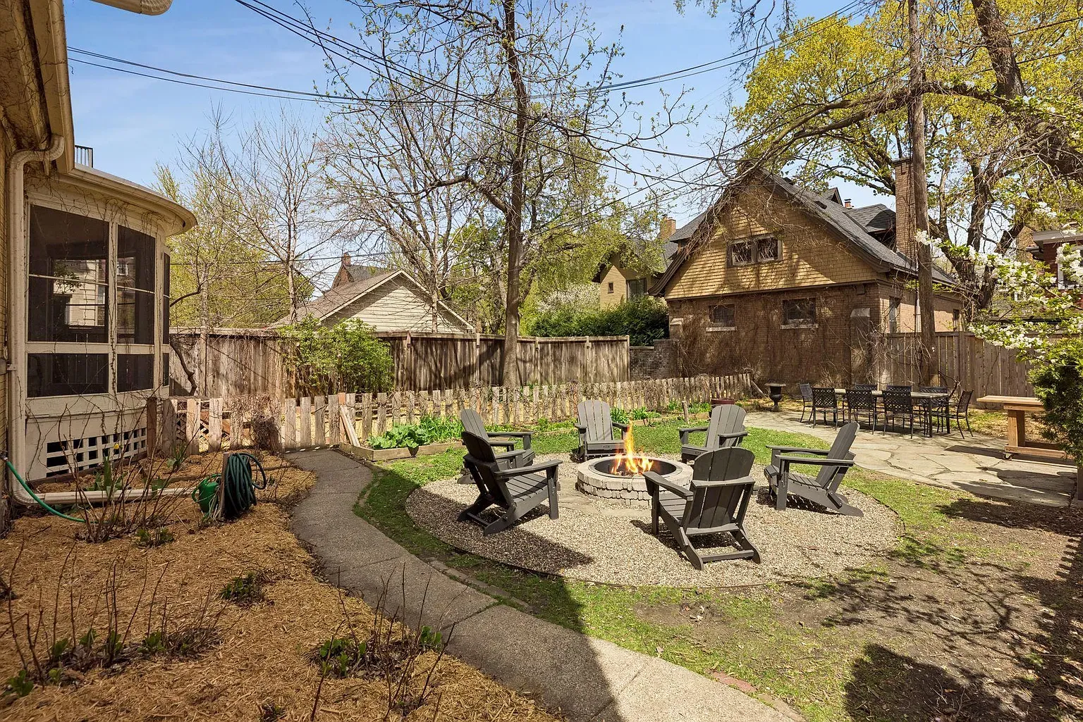 Backyard scene with a circular stone fire pit surrounded by Adirondack chairs on gravel, a wooden fence, and leafless trees under a blue sky. Left shows a screened porch; right has a patio table and chairs.