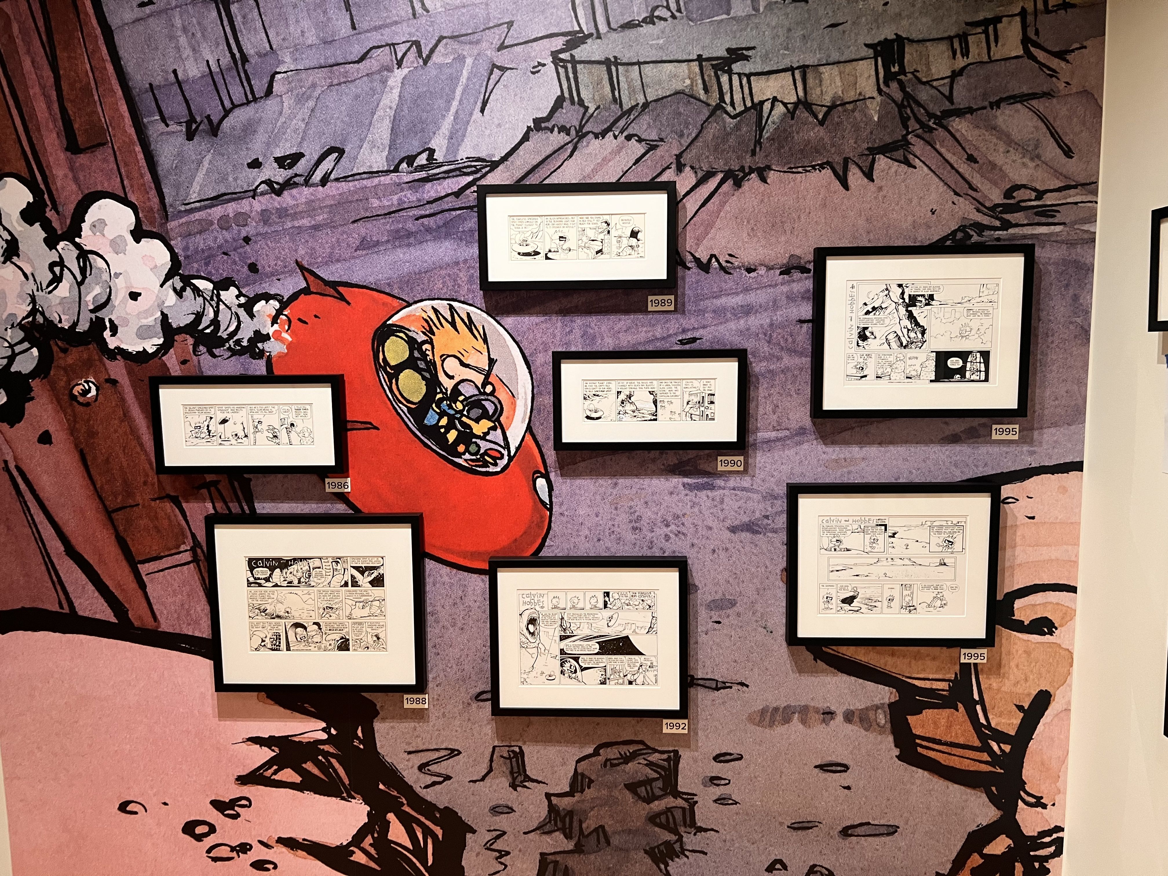 Purple wall mural featuring a large red space helmet and several framed Calvin and Hobbes comic strips (1986–1995) arranged in a grid.